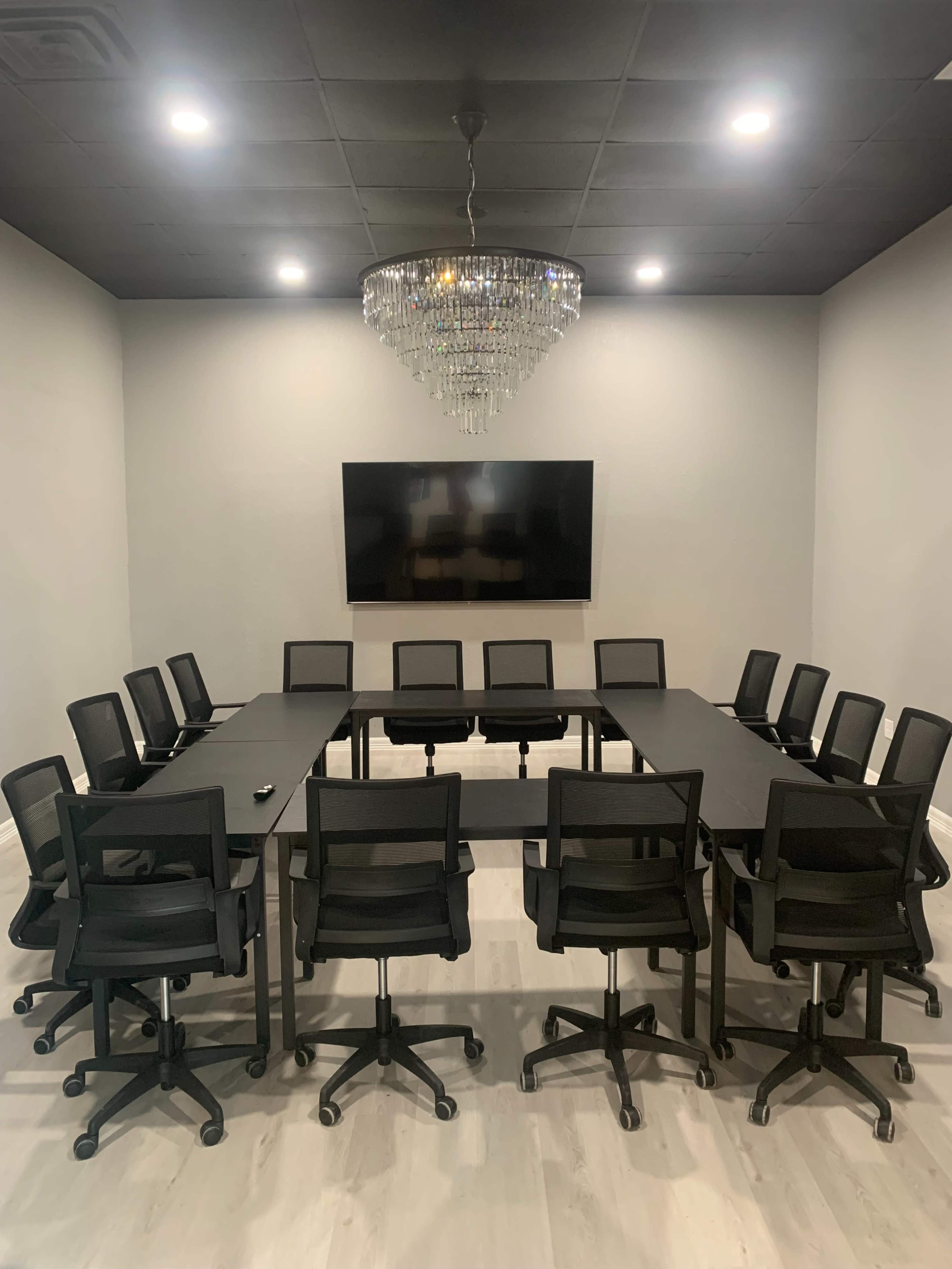 A conference room features a large rectangular table surrounded by black rolling chairs, with a chandelier overhead and a wall-mounted television.