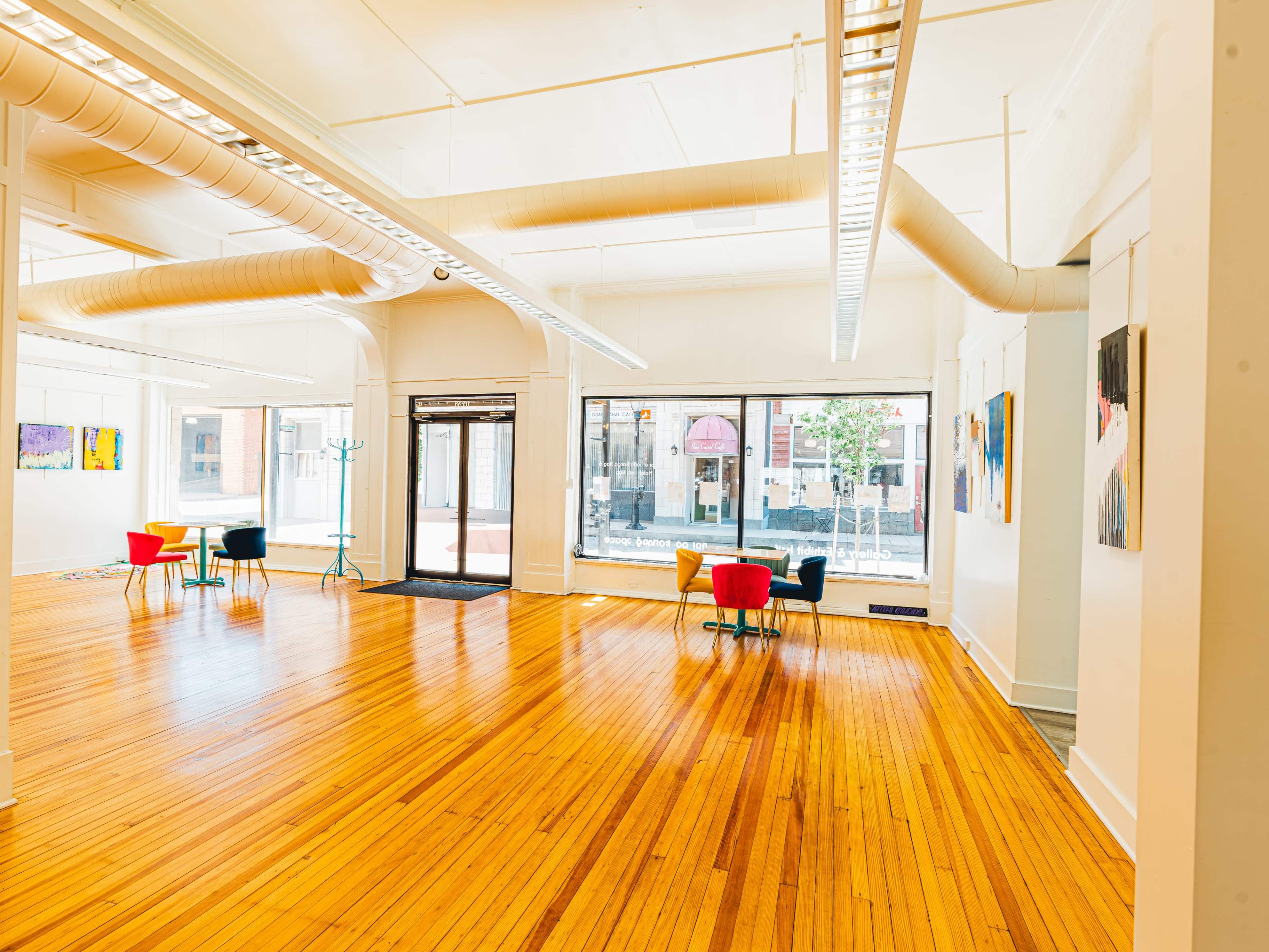 The image shows a bright, spacious room with polished wooden floors, large windows, and a few colorful chairs arranged near the entrance.