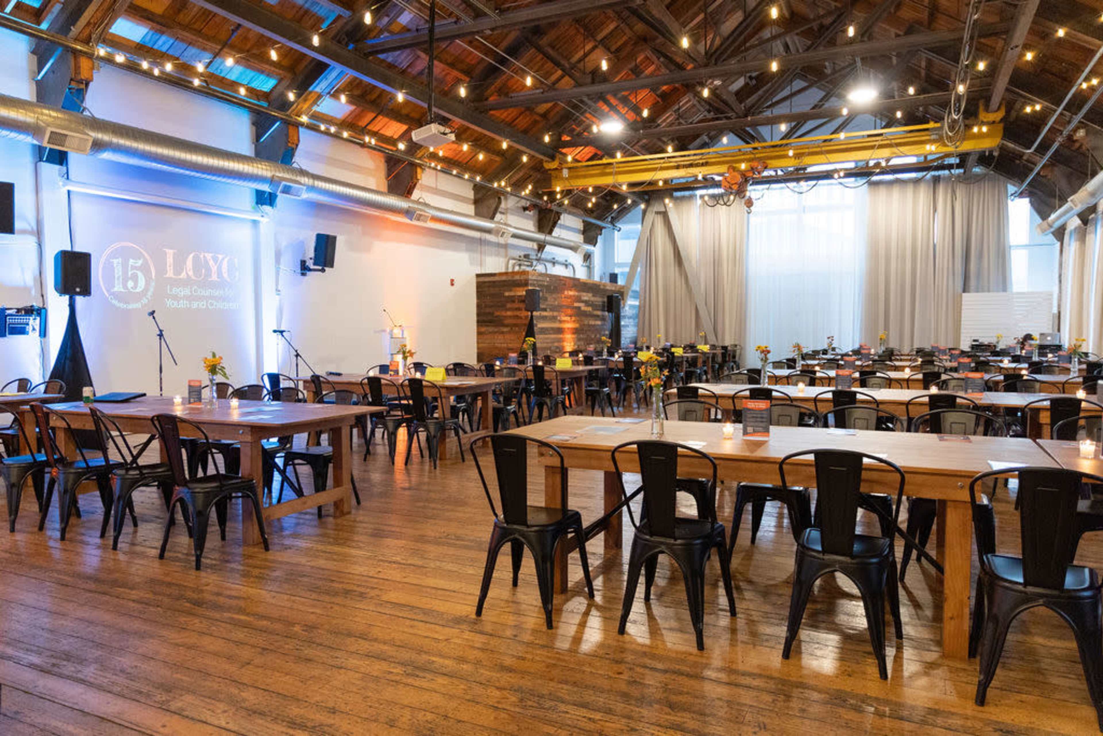 The image shows an inviting event space with wooden floors, tables set for dining, and a stage area with lighting and sound equipment.