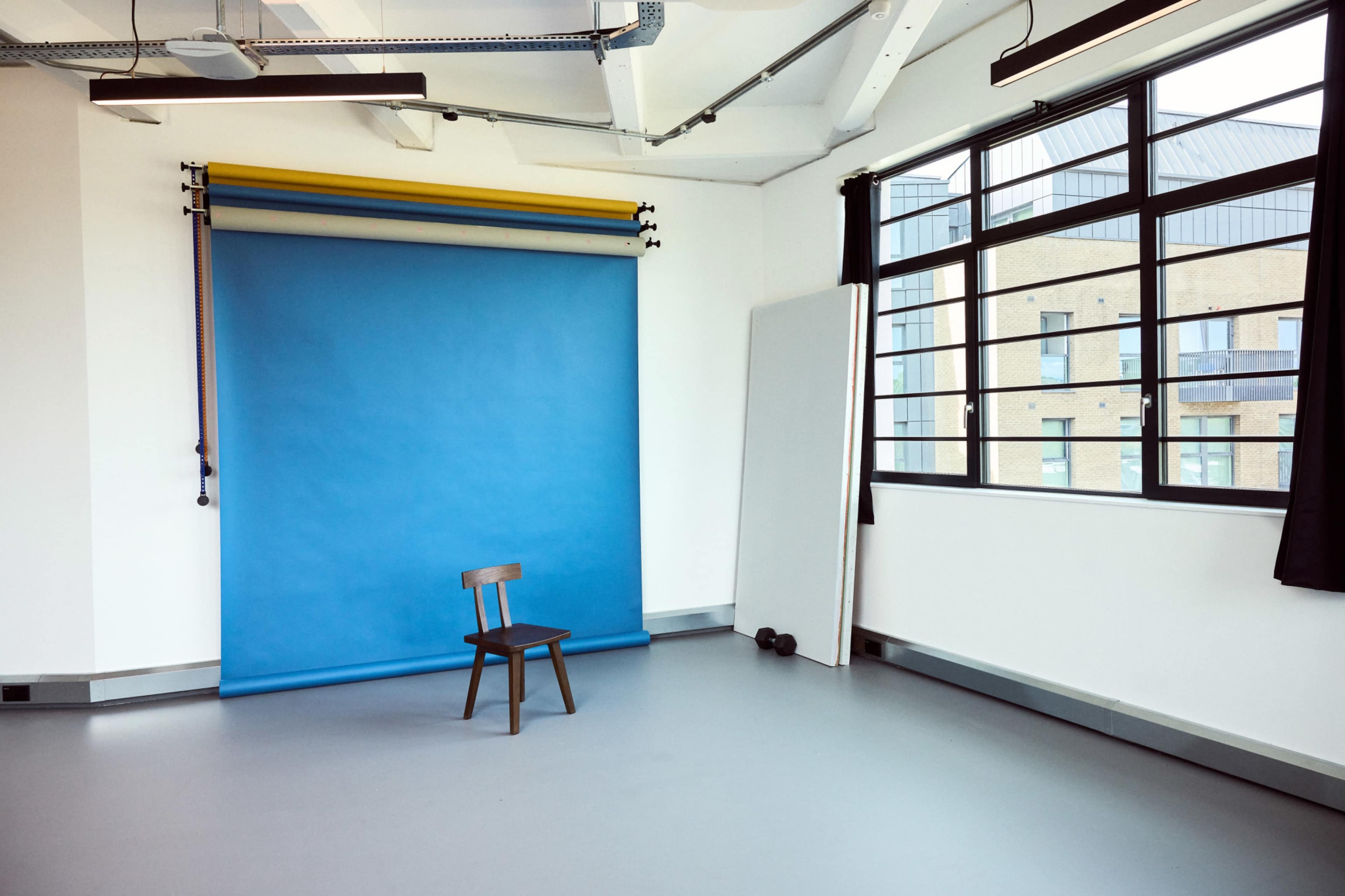 The image shows a simple studio space with a blue backdrop, a wooden chair, a large white panel, and a pair of dumbbells near the window.