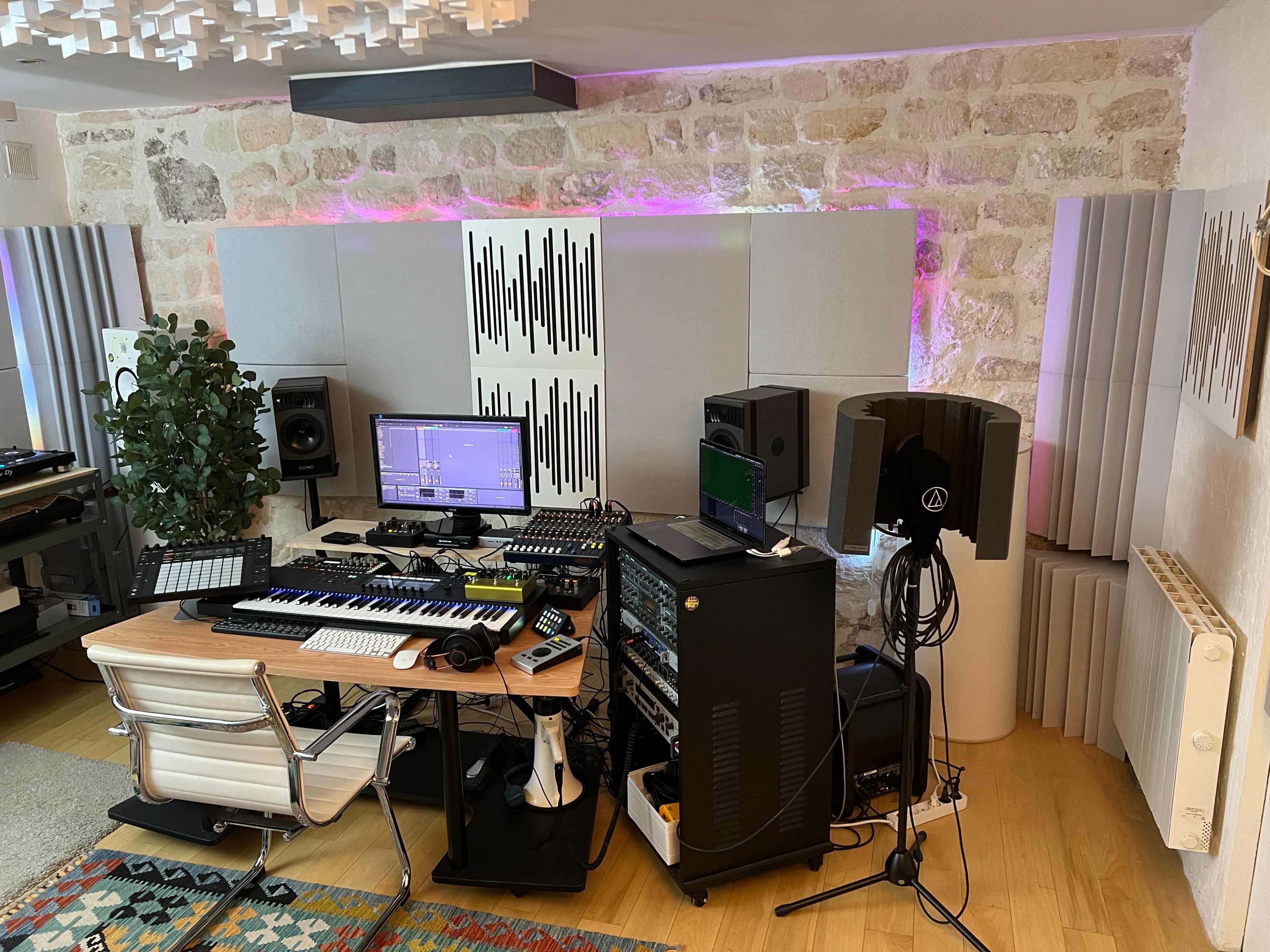 A modern music production studio features a desk with musical equipment, monitors, and a computer setup against a backdrop of stone walls and acoustic panels.