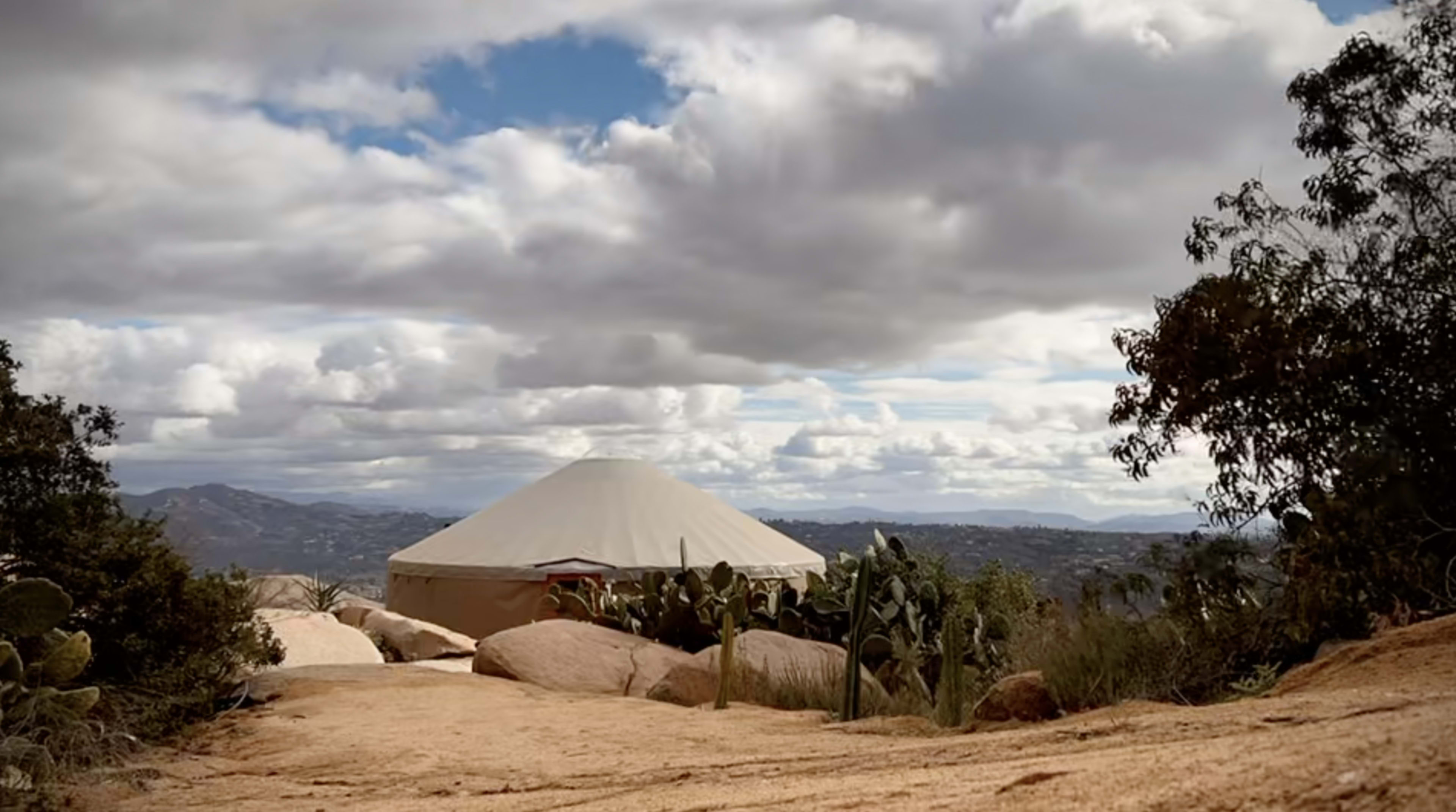 A yurt stands on a rocky path surrounded by cacti and a view of distant hills under a cloudy sky.