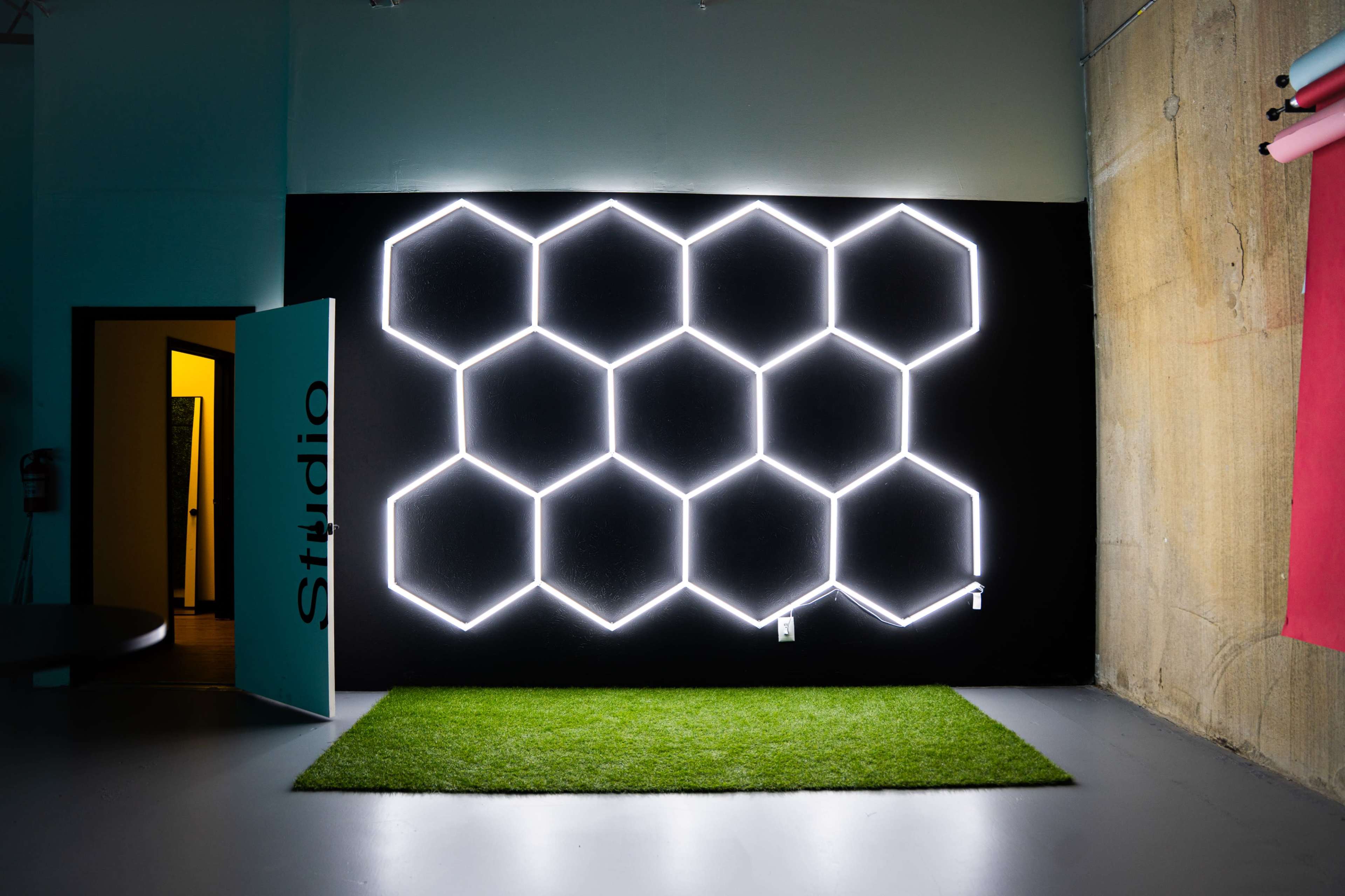 The image shows a black wall with a hexagonal light installation, next to a green artificial grass area and an open door leading to a room labeled "Studio."
