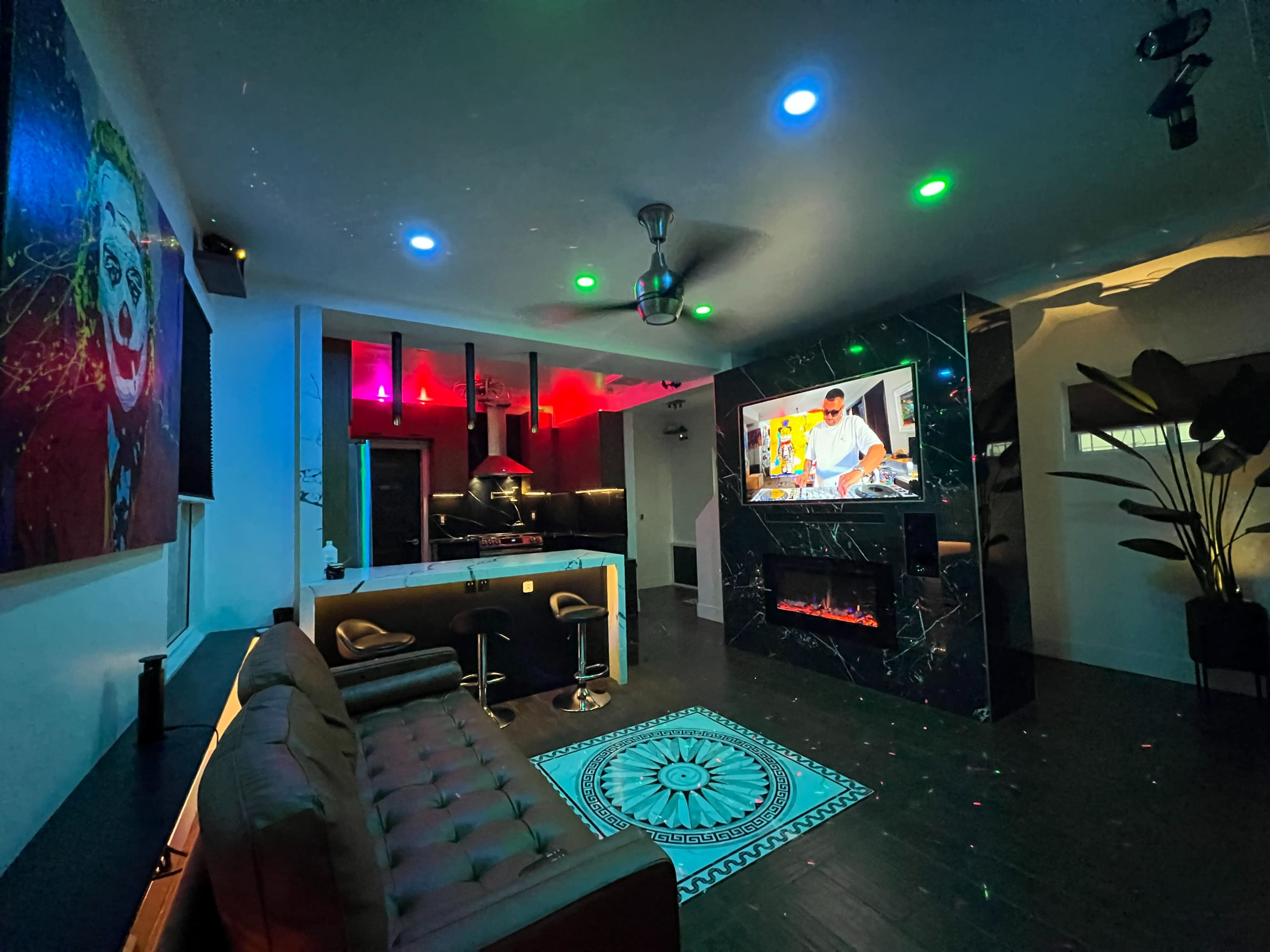 A modern living space features a sofa, a bar area, and a large television displaying a DJ, with colorful lights illuminating the room.
