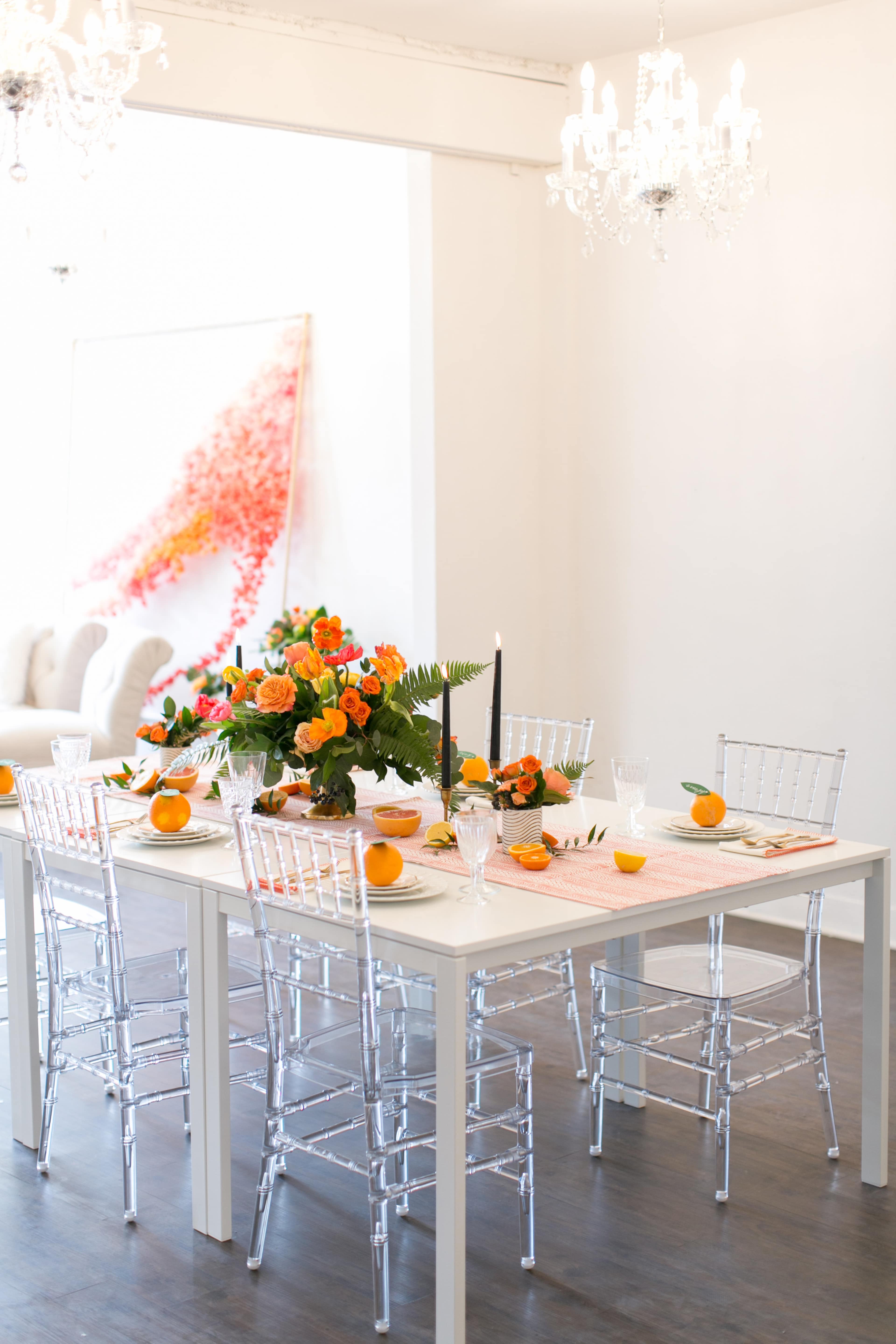 A dining table is set with a colorful floral centerpiece and oranges, surrounded by clear acrylic chairs in a well-lit room.