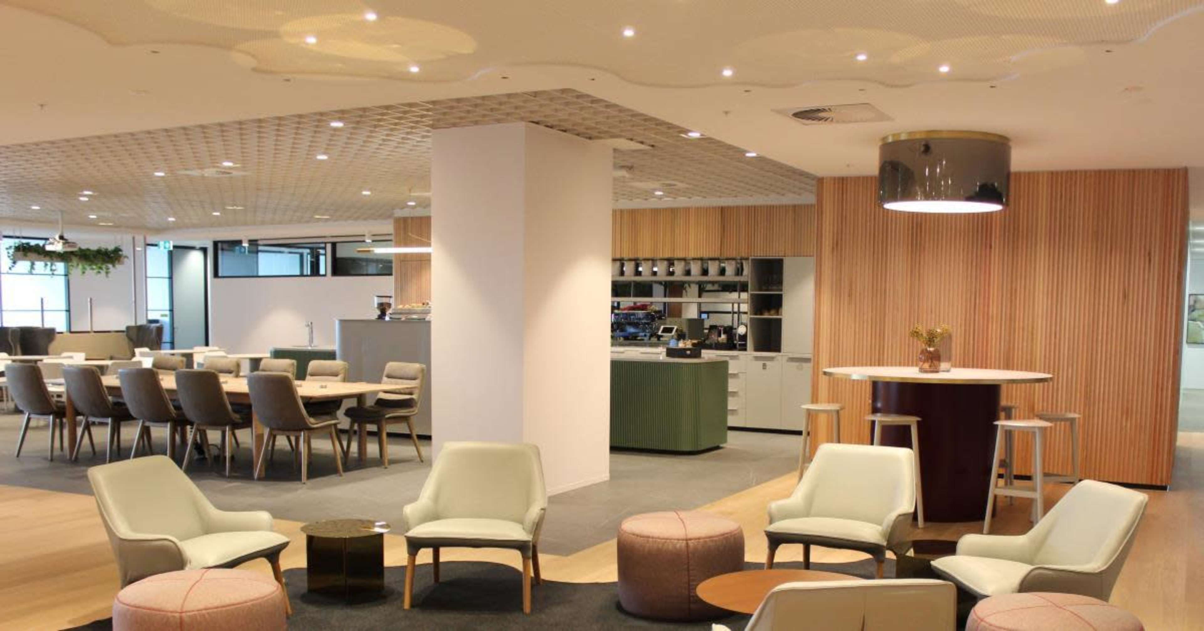 The image shows a modern office interior featuring a dining area with multiple seating arrangements, a coffee bar, and wooden accents on the walls.