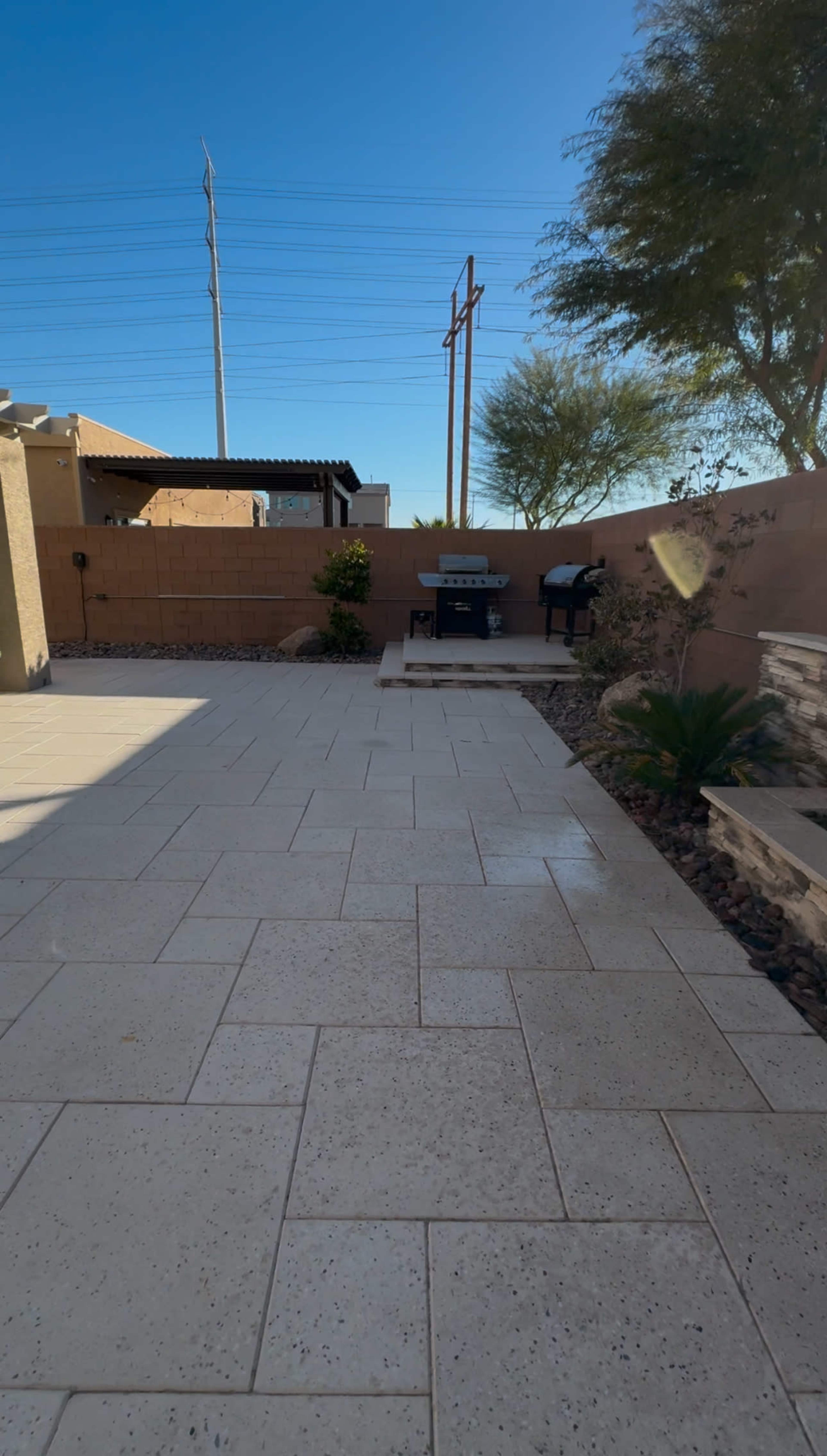 The image shows a paved backyard space with a barbecue grill, plantings along the walls, and clear blue skies above.