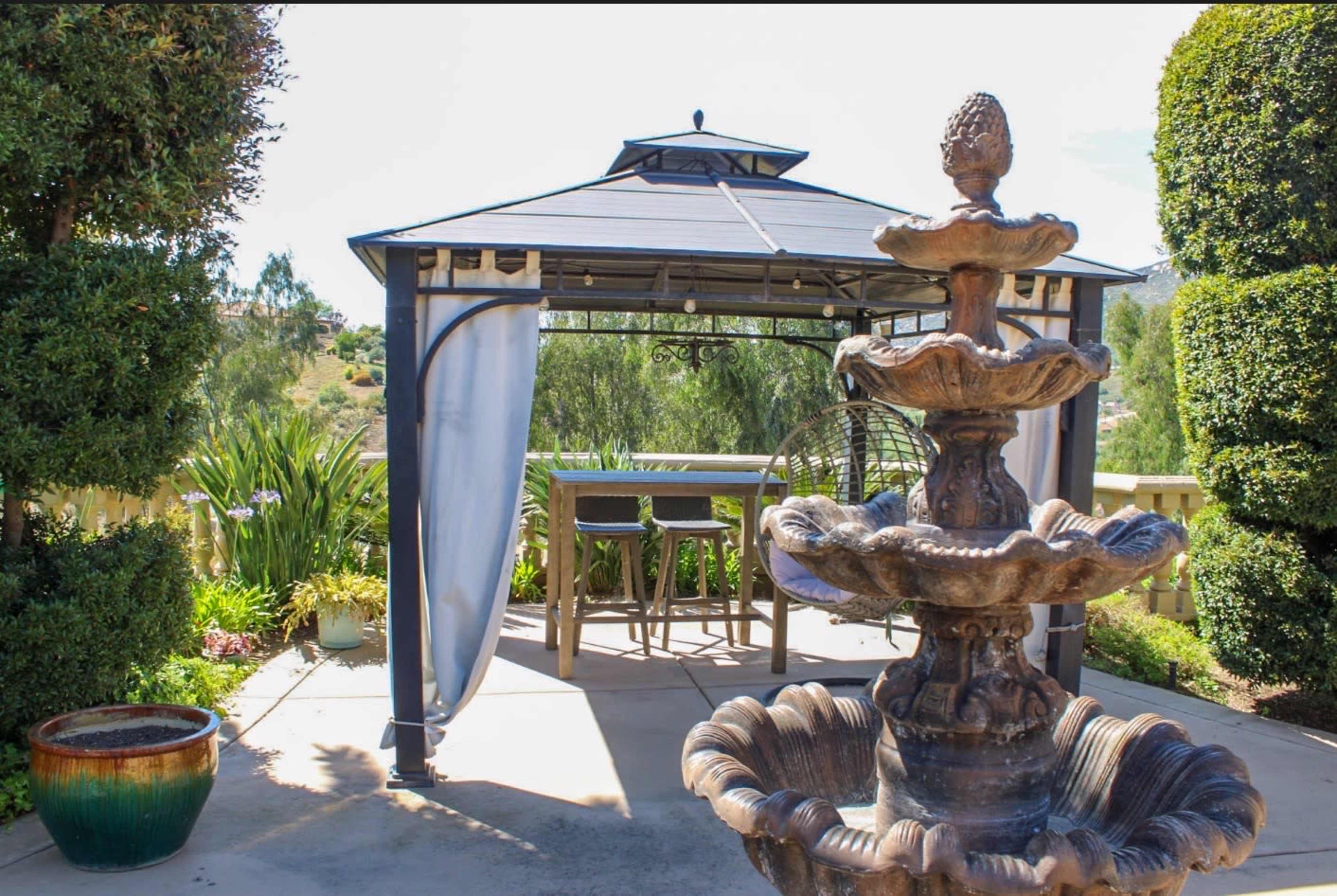 A gazebo with a fountain and seating area is situated in a landscaped garden.