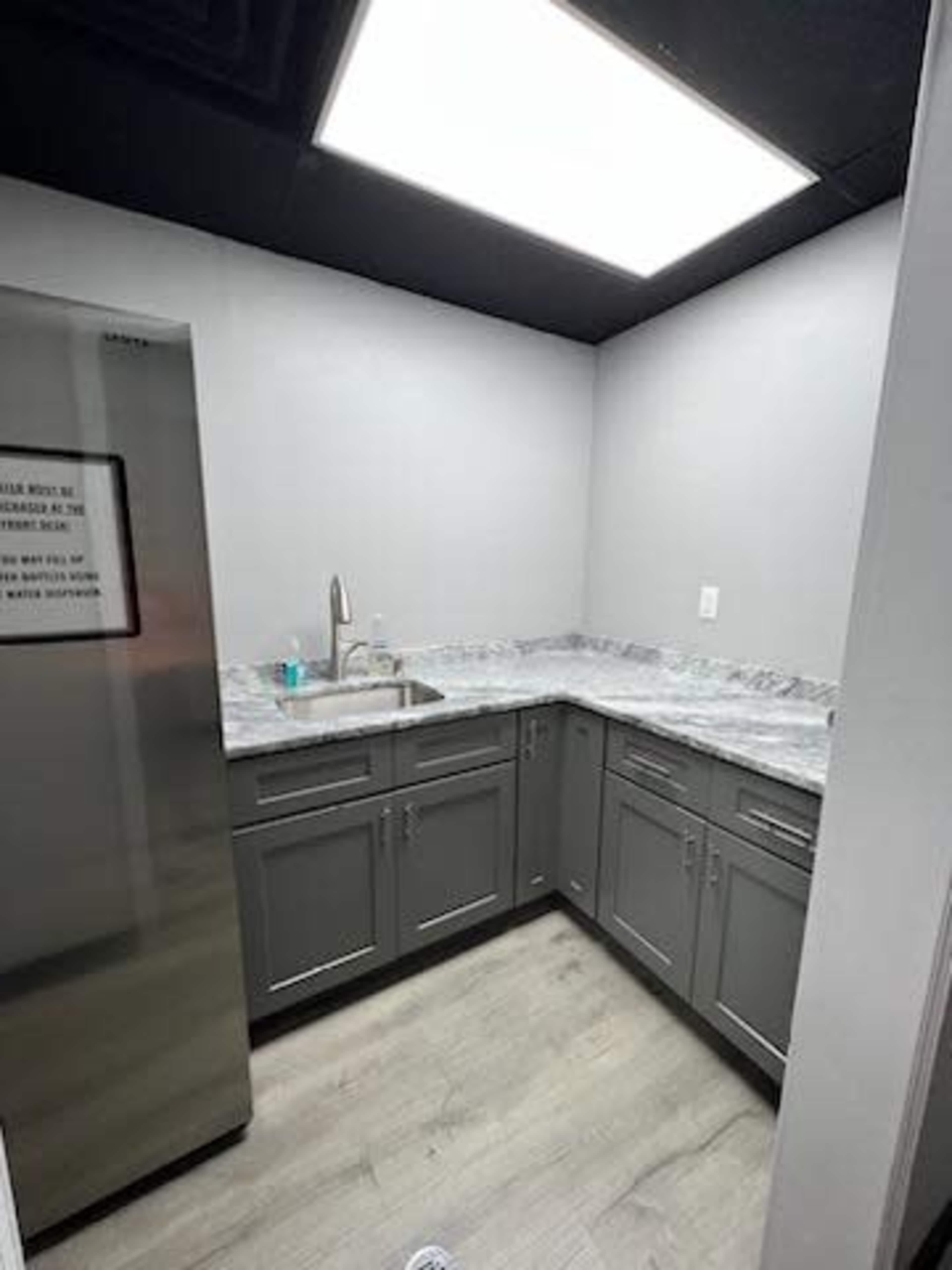 The image shows a small kitchen with gray cabinetry, a countertop made of gray stone, a stainless steel refrigerator, and a sink under a bright overhead light.