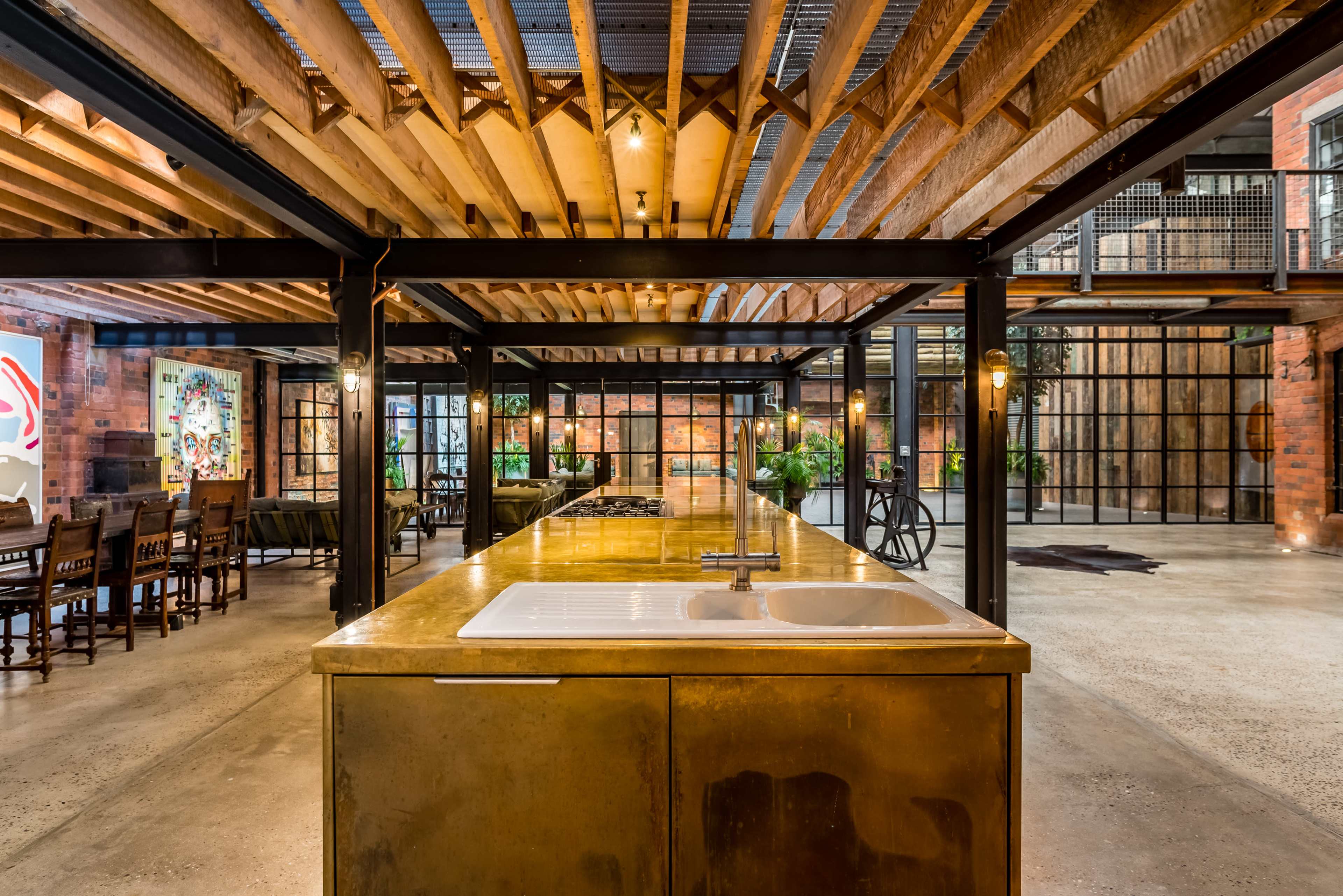 A modern industrial kitchen with a large metal countertop and exposed wooden ceiling beams, surrounded by a spacious dining area with brick walls and large windows.