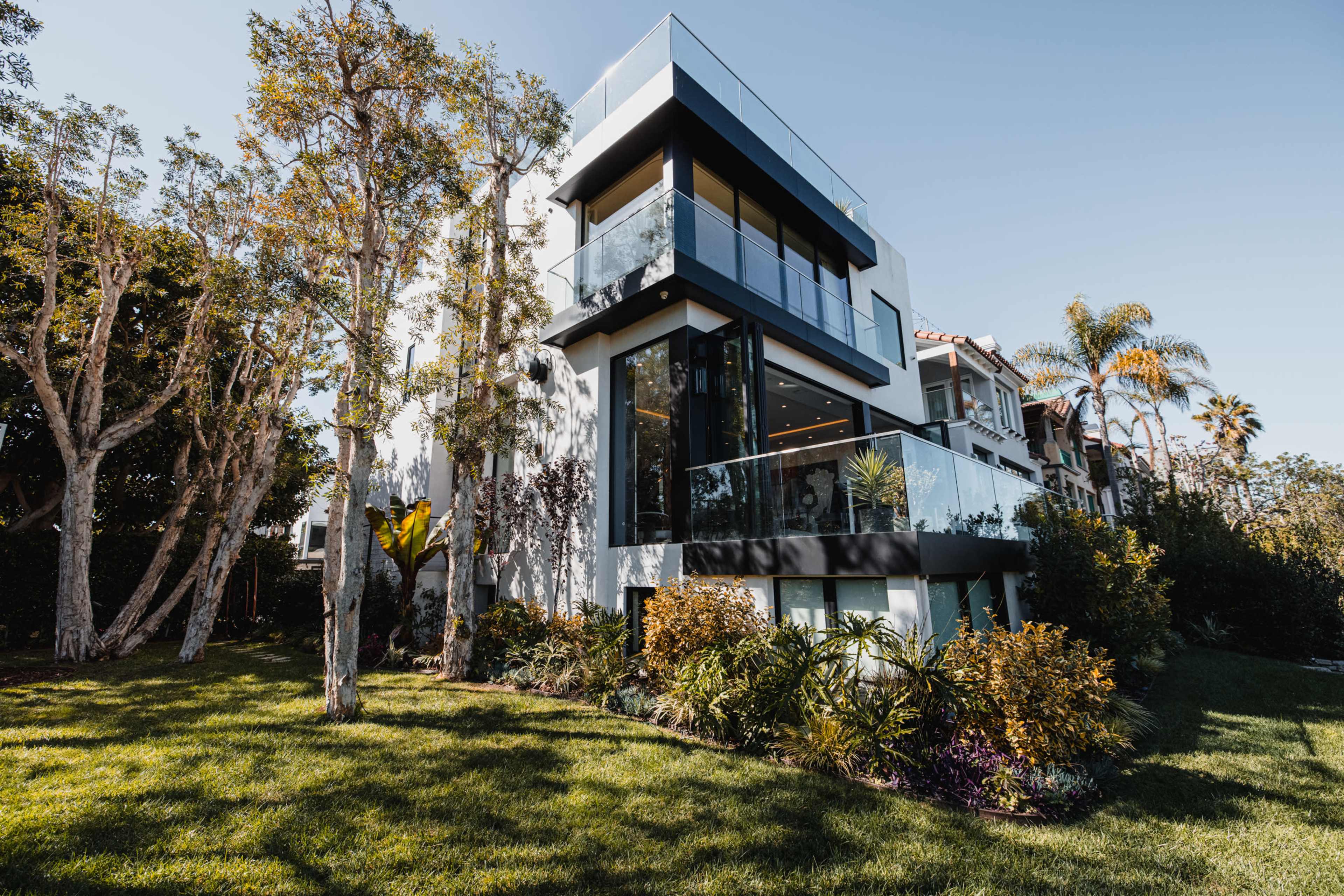 A modern, multi-story house with large glass balconies is surrounded by lush greenery and palm trees.