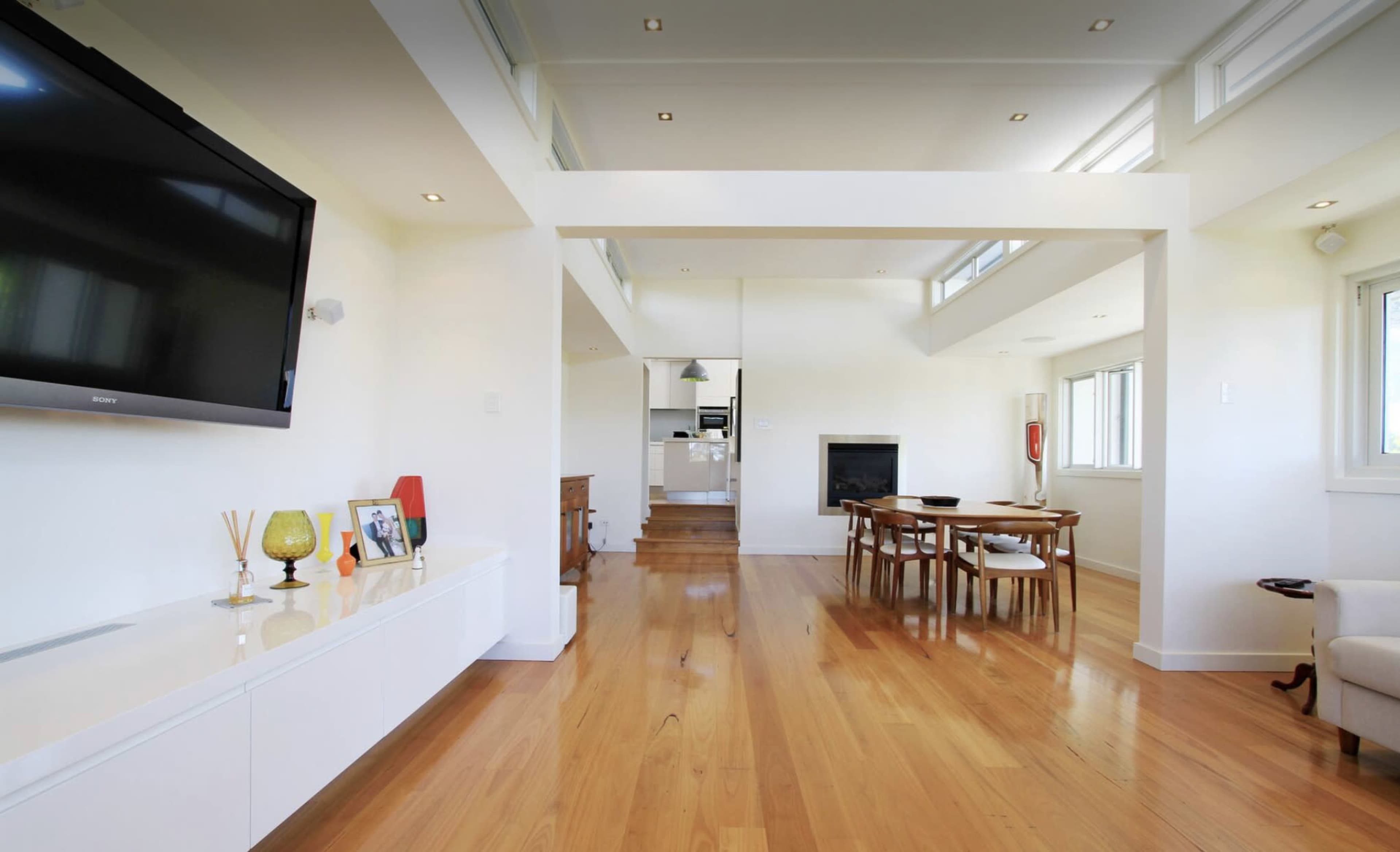 The image shows a modern living space with wooden flooring, a television mounted on the wall, and a dining area with a table and chairs.