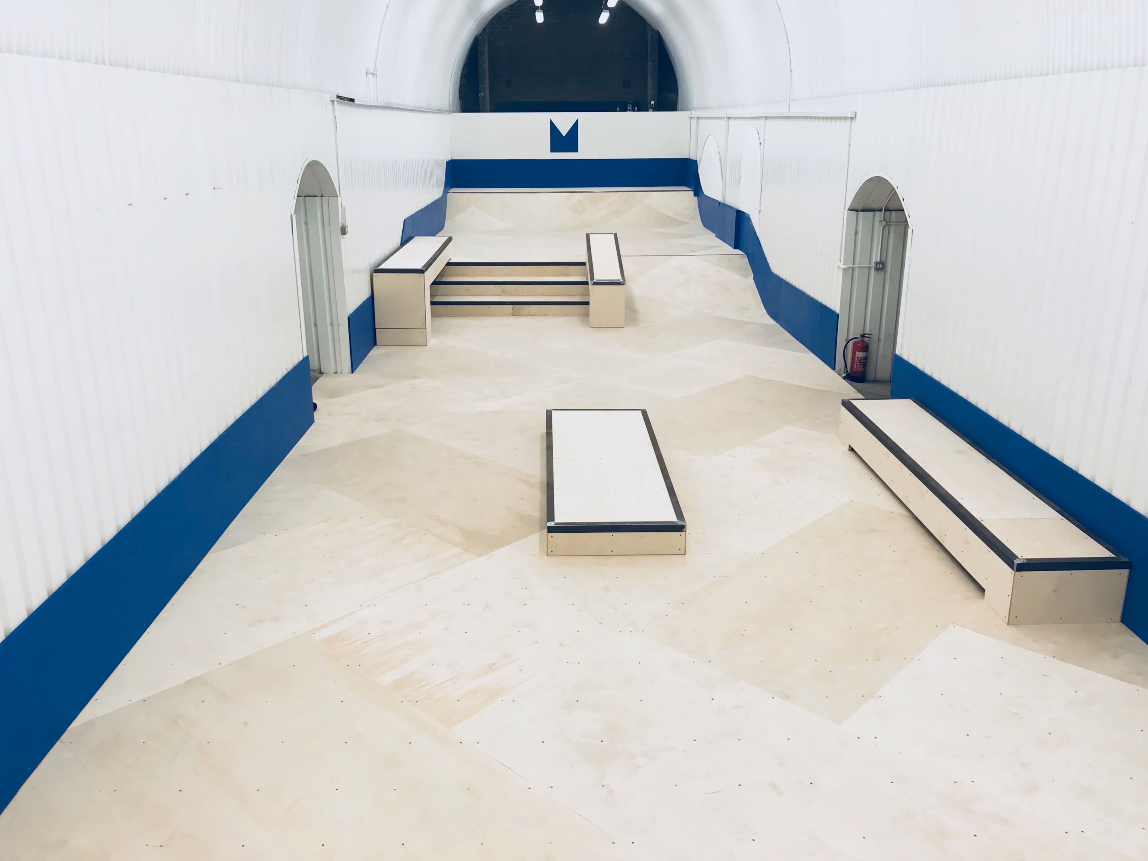 A skatepark interior featuring multiple ramps and flat surfaces arranged in a tunnel-like structure with white walls and blue accents.