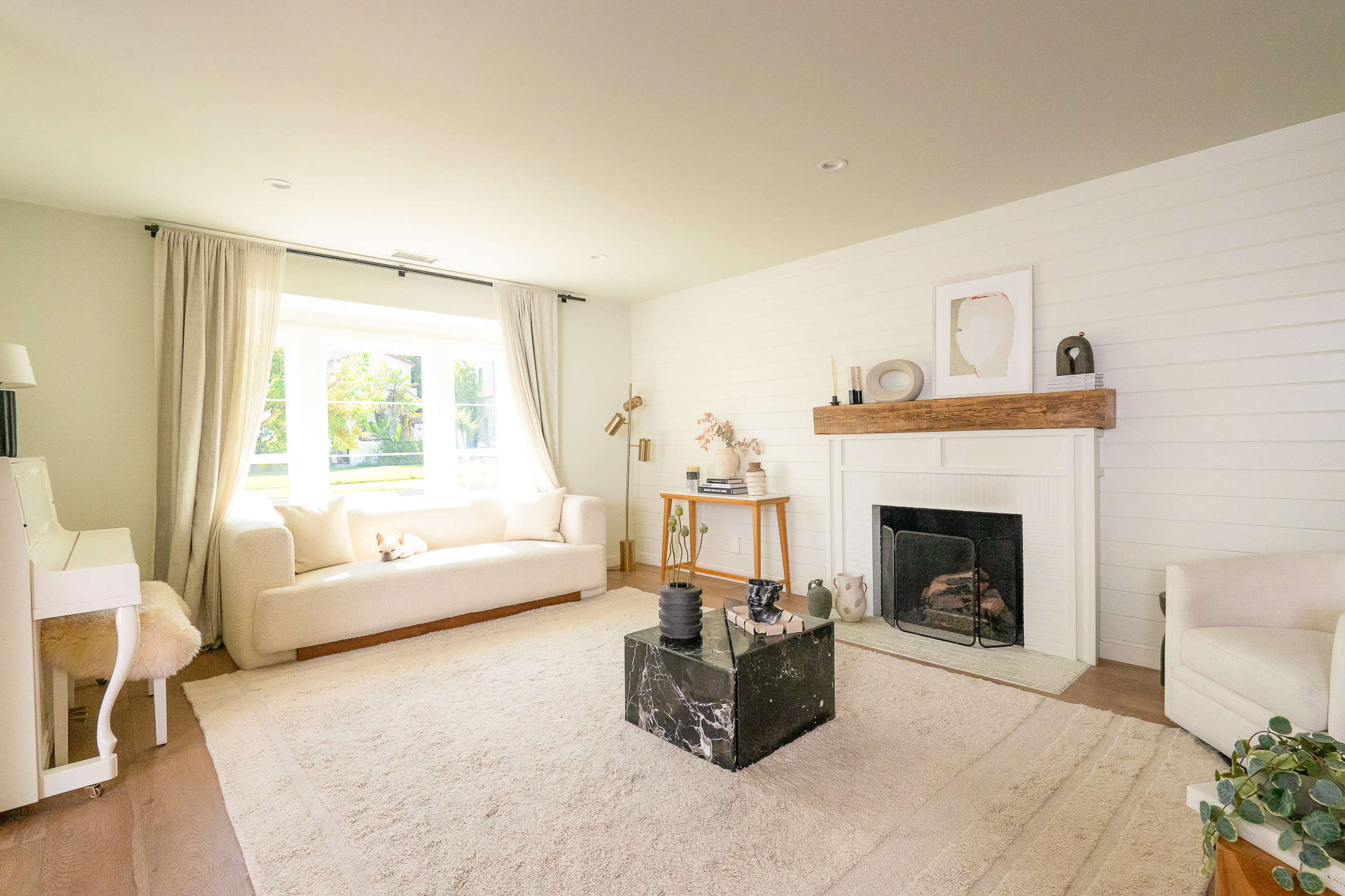 The living room features a white shiplap wall, a fireplace with a wooden mantel, a plush rug, and large windows allowing natural light to fill the space.