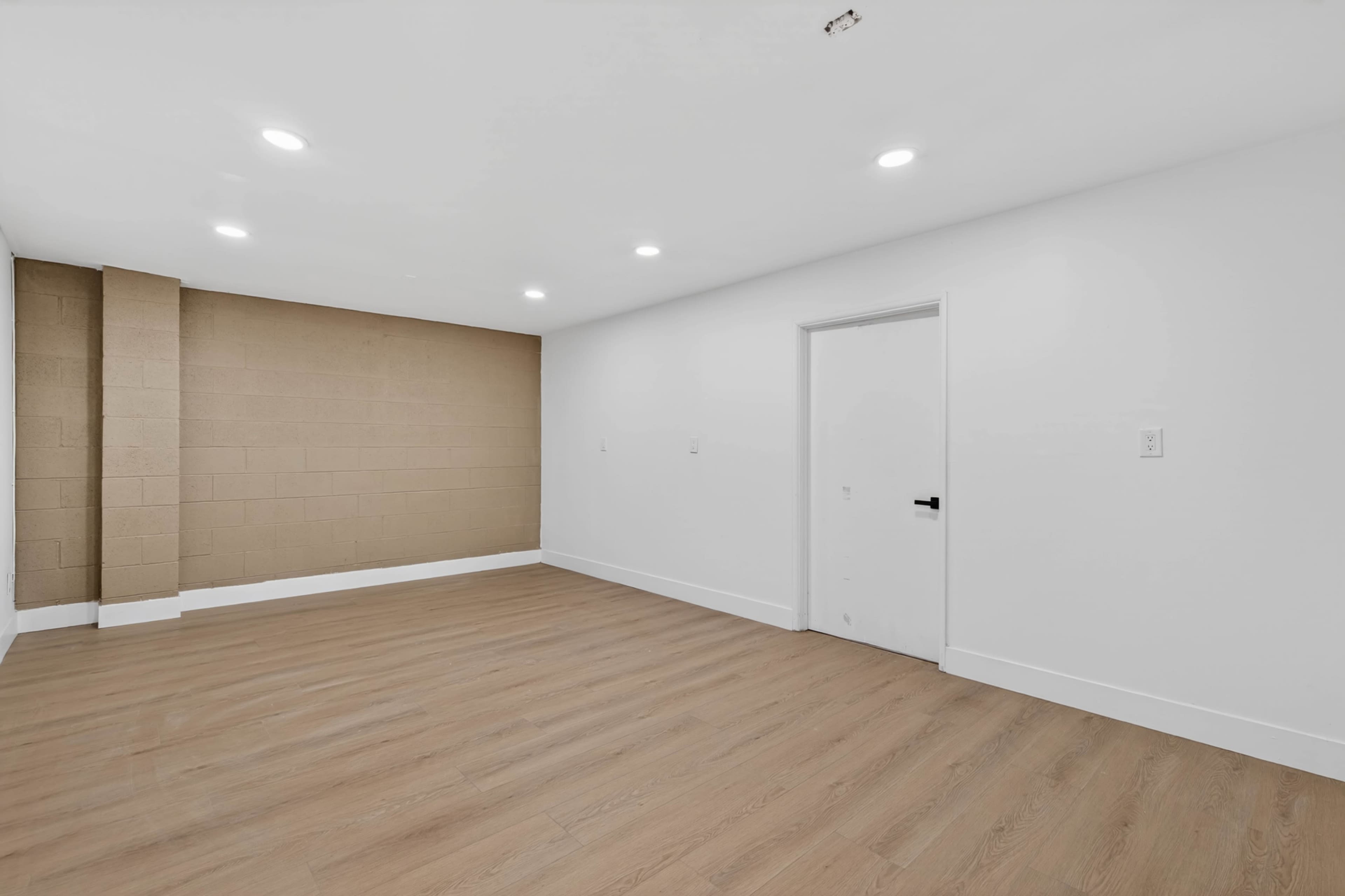 The image shows an empty room with a light wooden floor, a light-colored wall, and a single door.