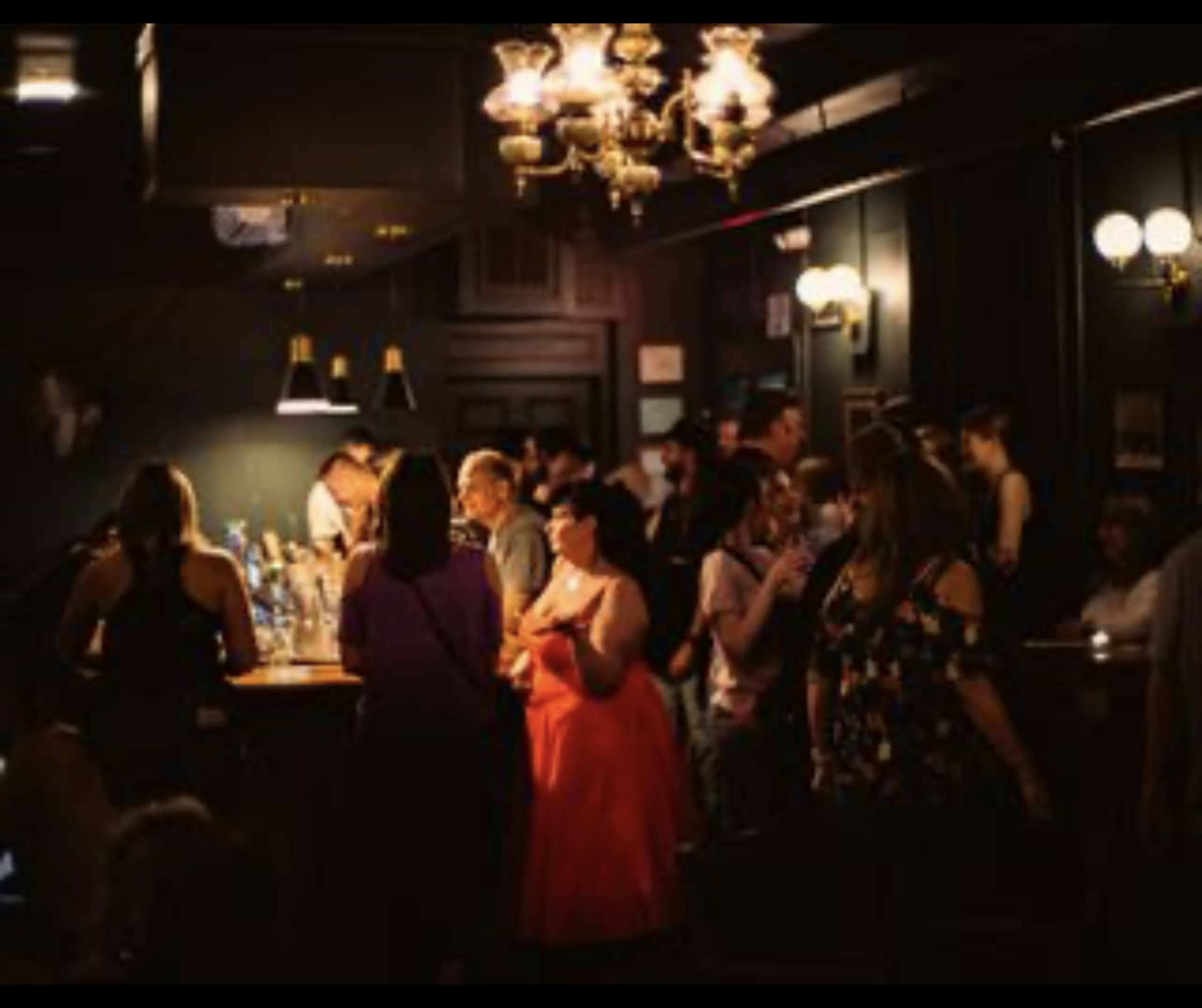 A crowded bar scene features patrons gathering around a well-lit bar while others socialize in a dimly lit environment.