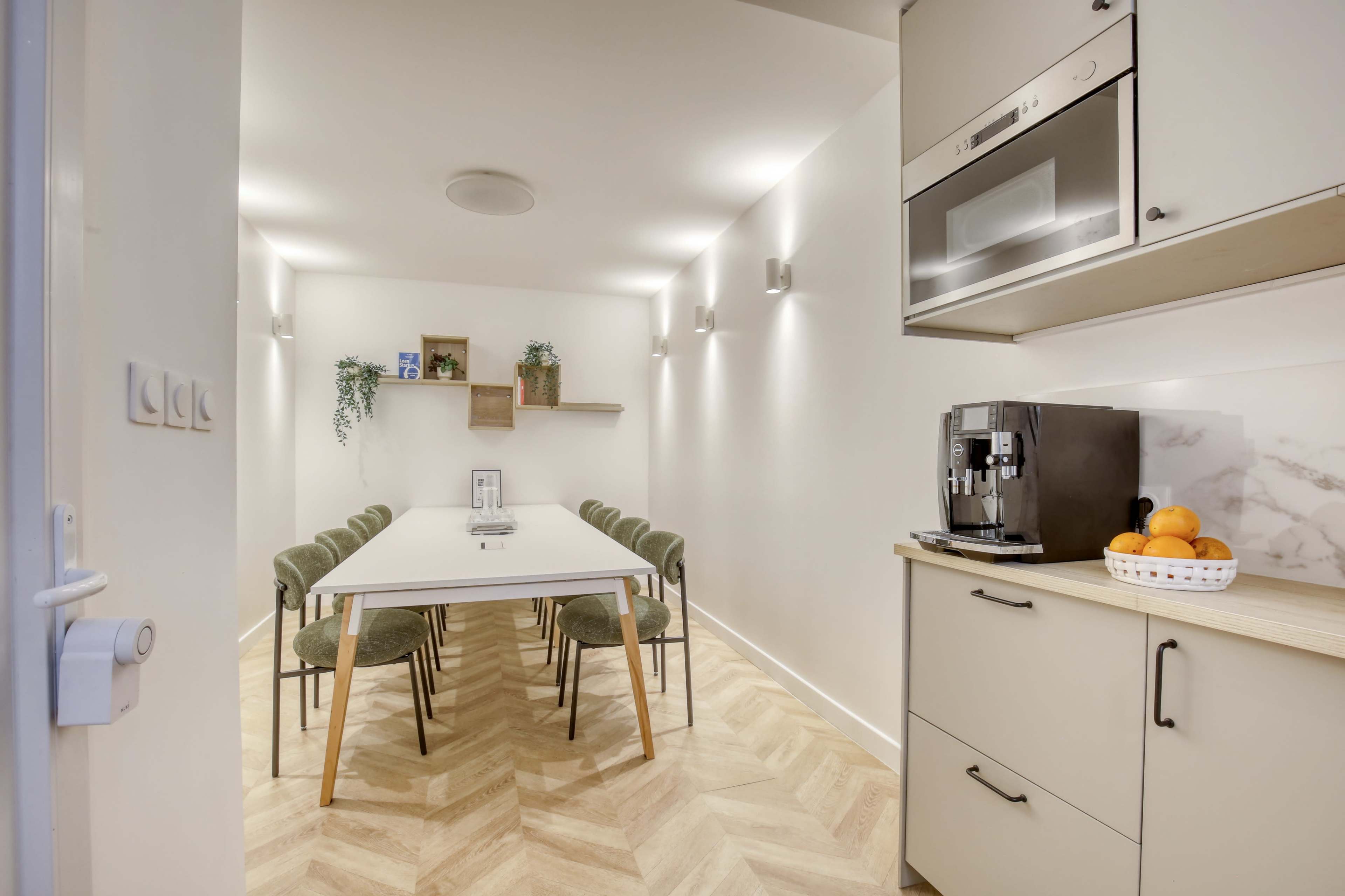 The image shows a modern, well-lit meeting room with a long rectangular table surrounded by green chairs, complemented by a small kitchenette in the corner.