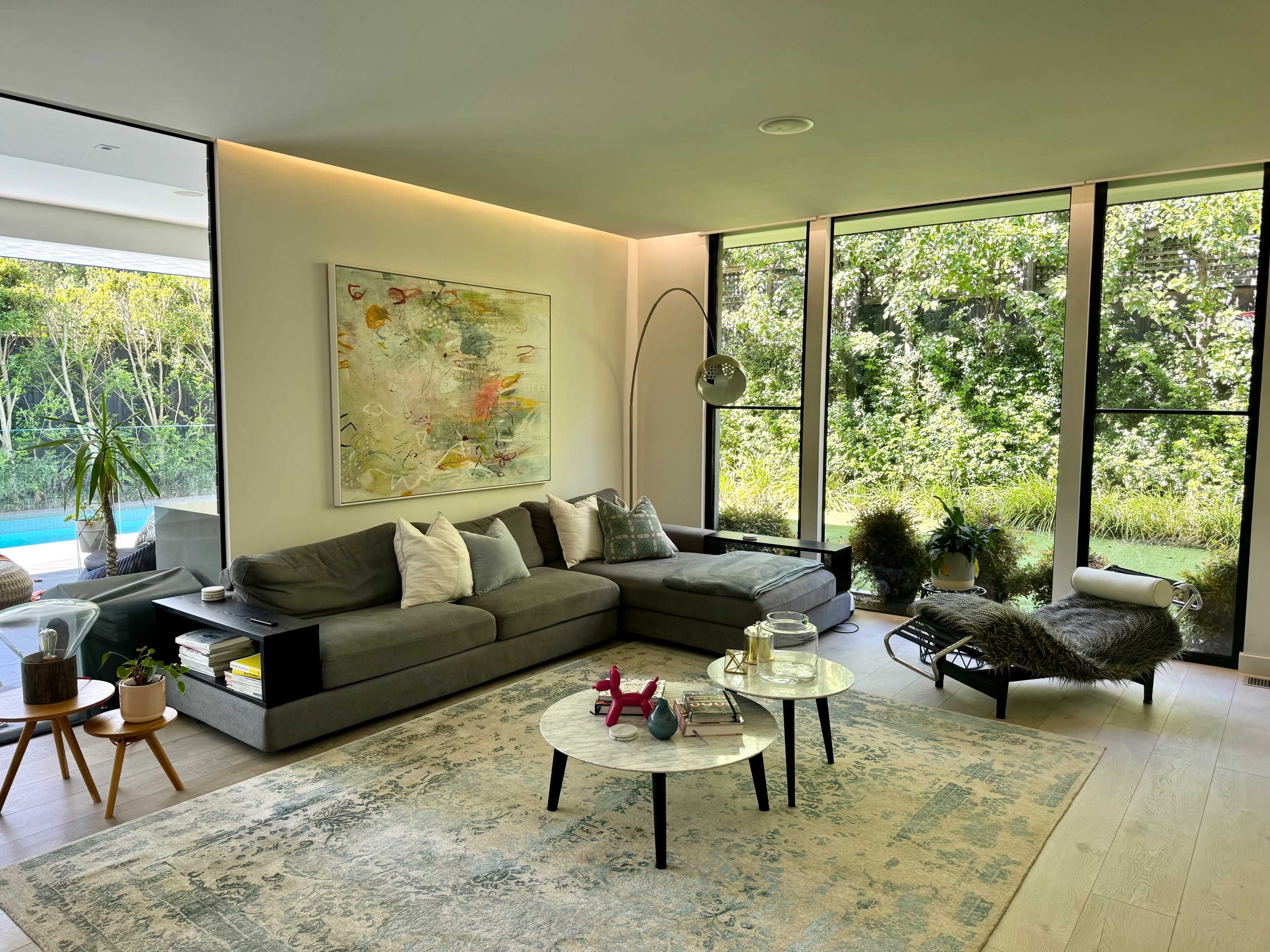 A modern living room features a large gray sectional sofa, a round coffee table, and floor-to-ceiling windows overlooking a green landscape.