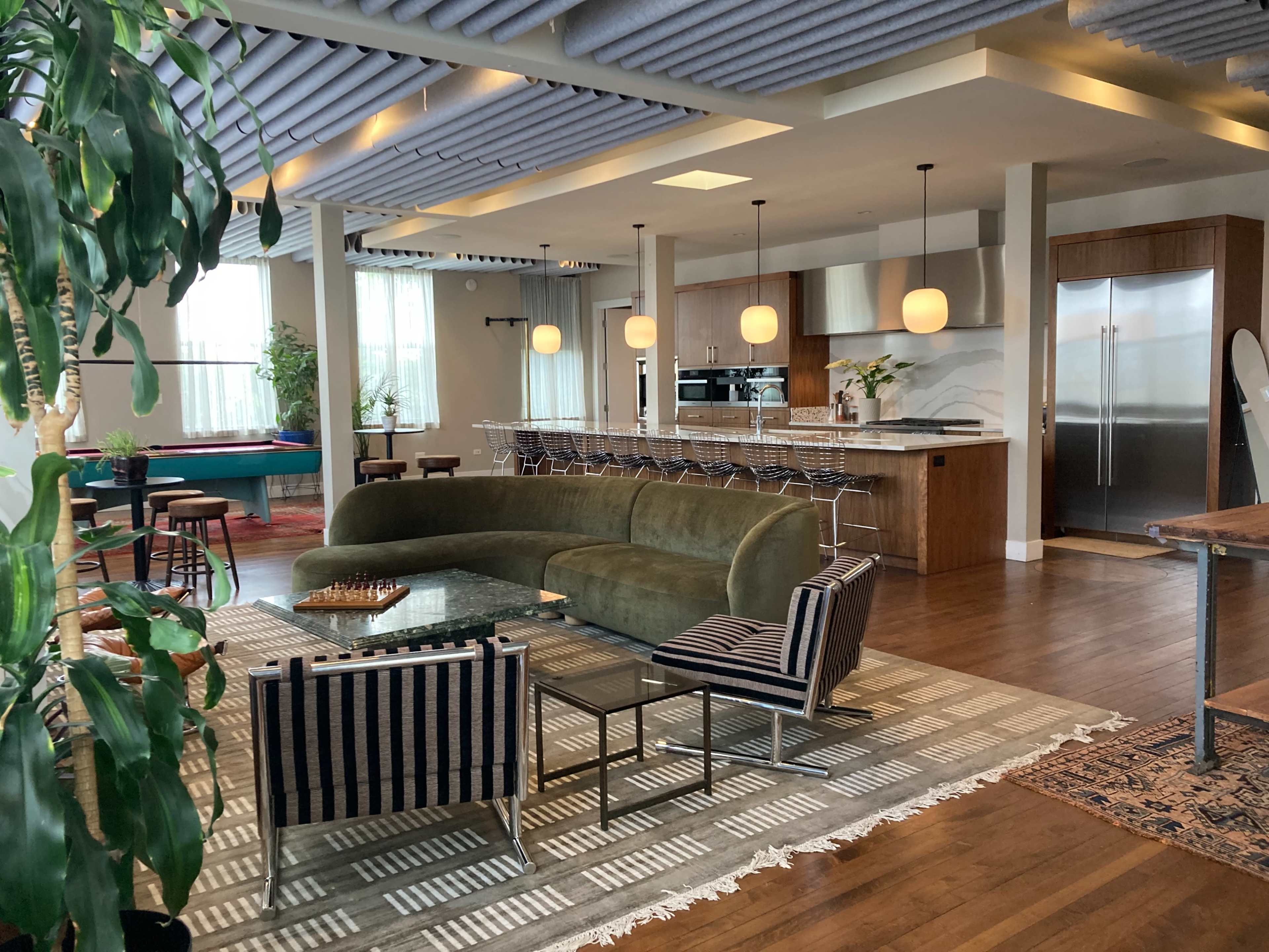 The image shows a modern open-concept living space featuring a curved green sofa, a kitchen with stainless steel appliances, and various seating areas.