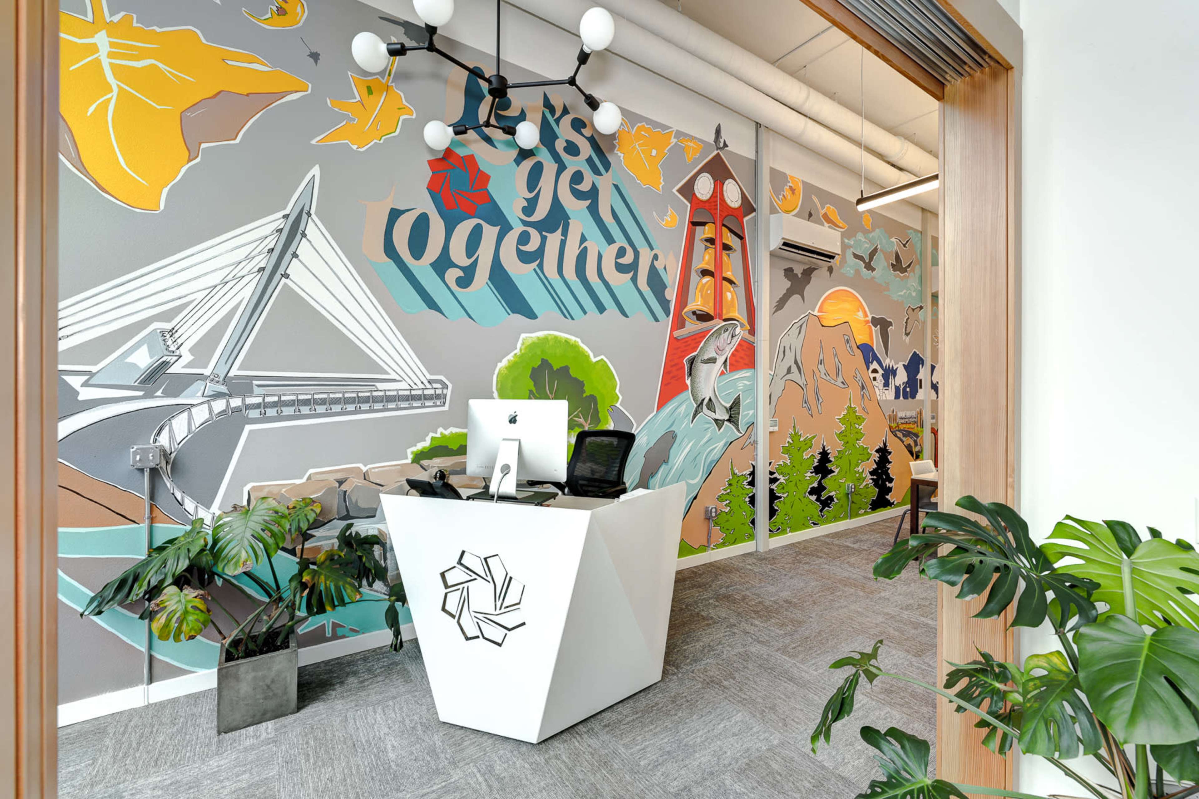 A vibrant office space with a large mural featuring a bridge and mountains, complemented by a modern white reception desk with a computer and potted plants.