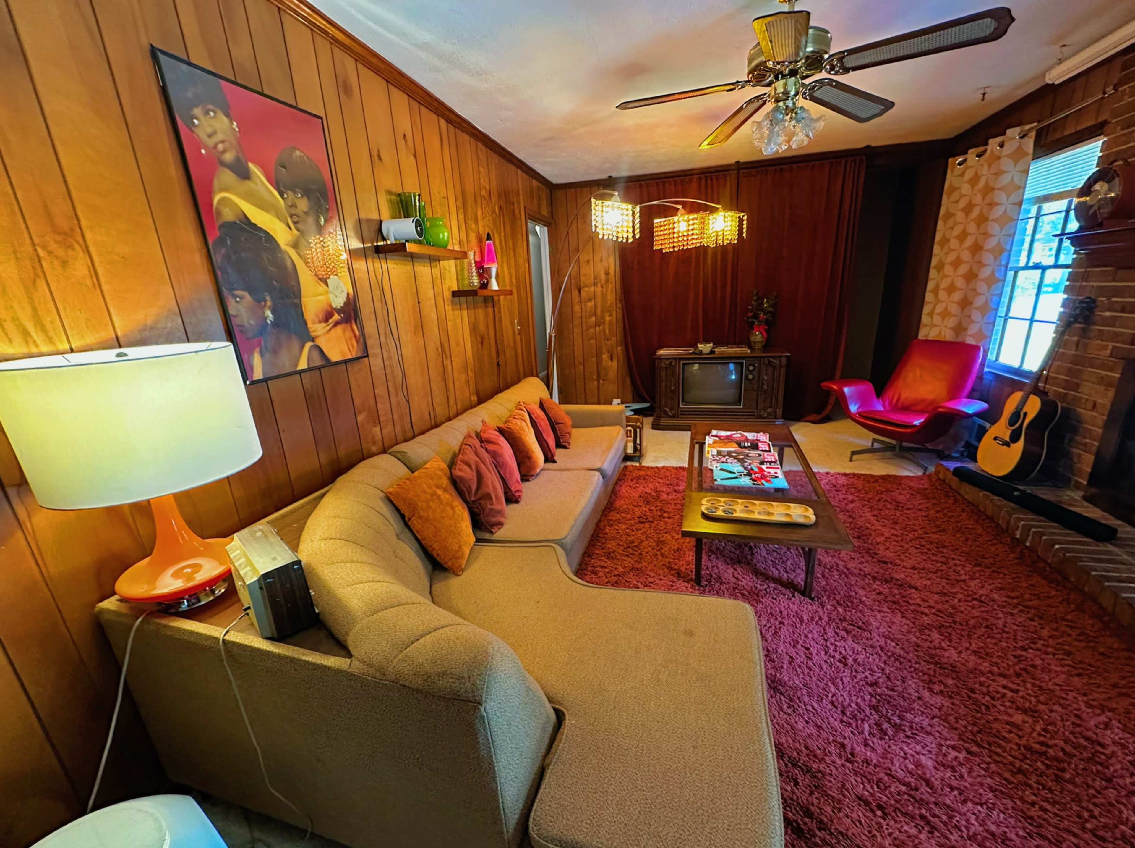 The living room features a sectional sofa with colorful cushions, a coffee table, a vintage television, and a guitar leaning against a brick wall, all illuminated by warm lighting.