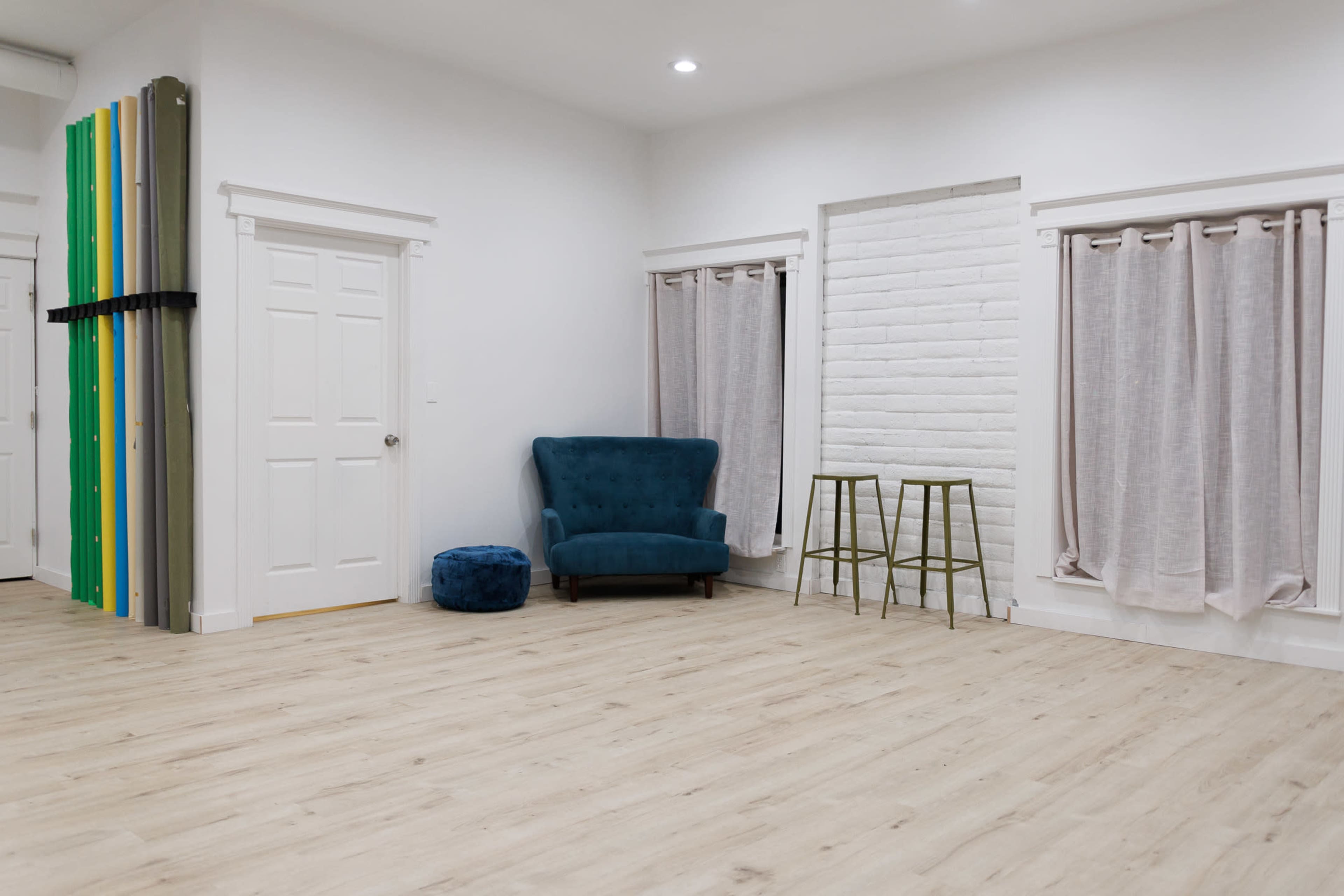The image shows a modern, empty room with light wood flooring, a blue armchair, a small blue pouf, two metal stools near a white brick wall, and curtains covering a window.