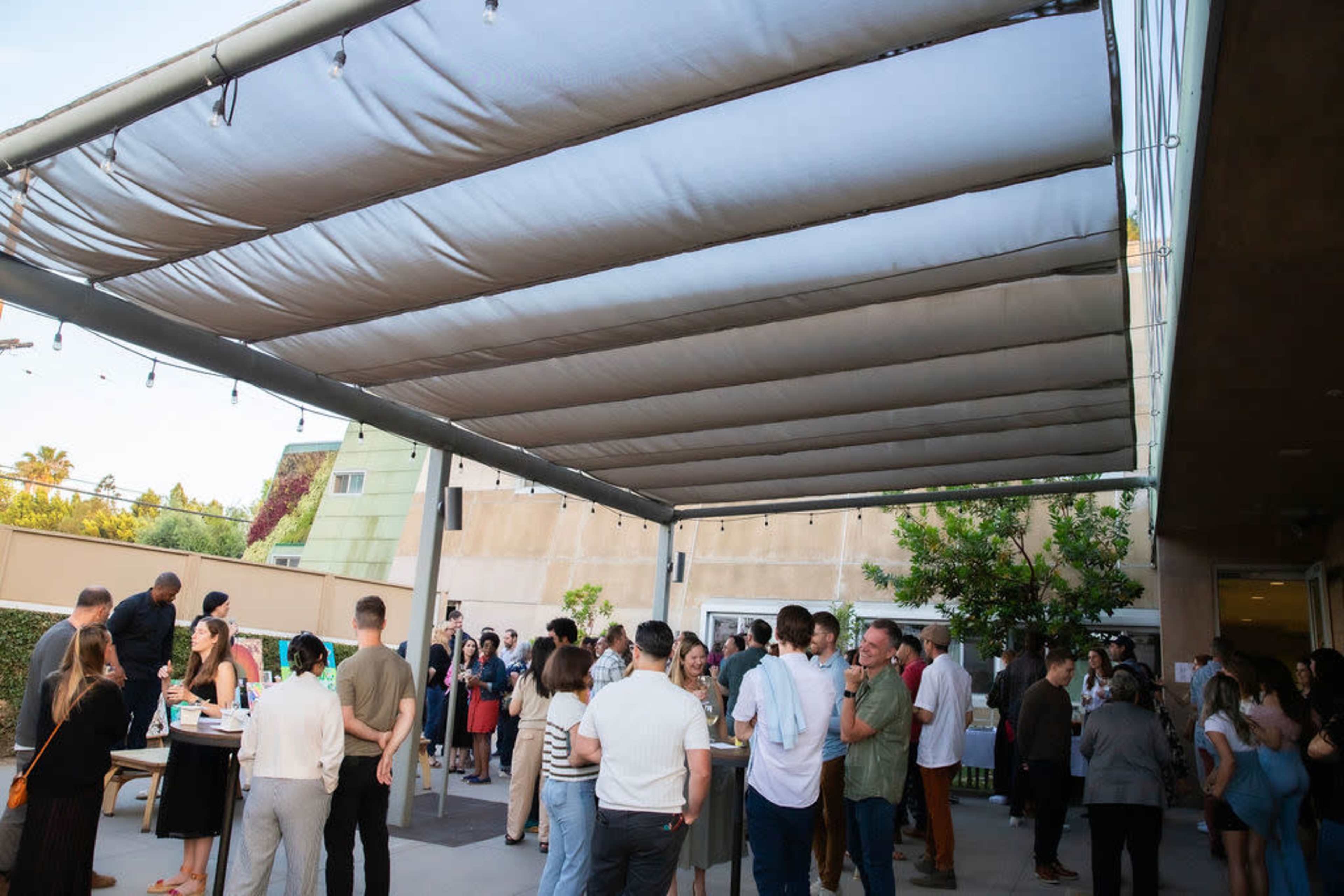 A large group of people mingles under a spacious, shaded canopy at an outdoor event.