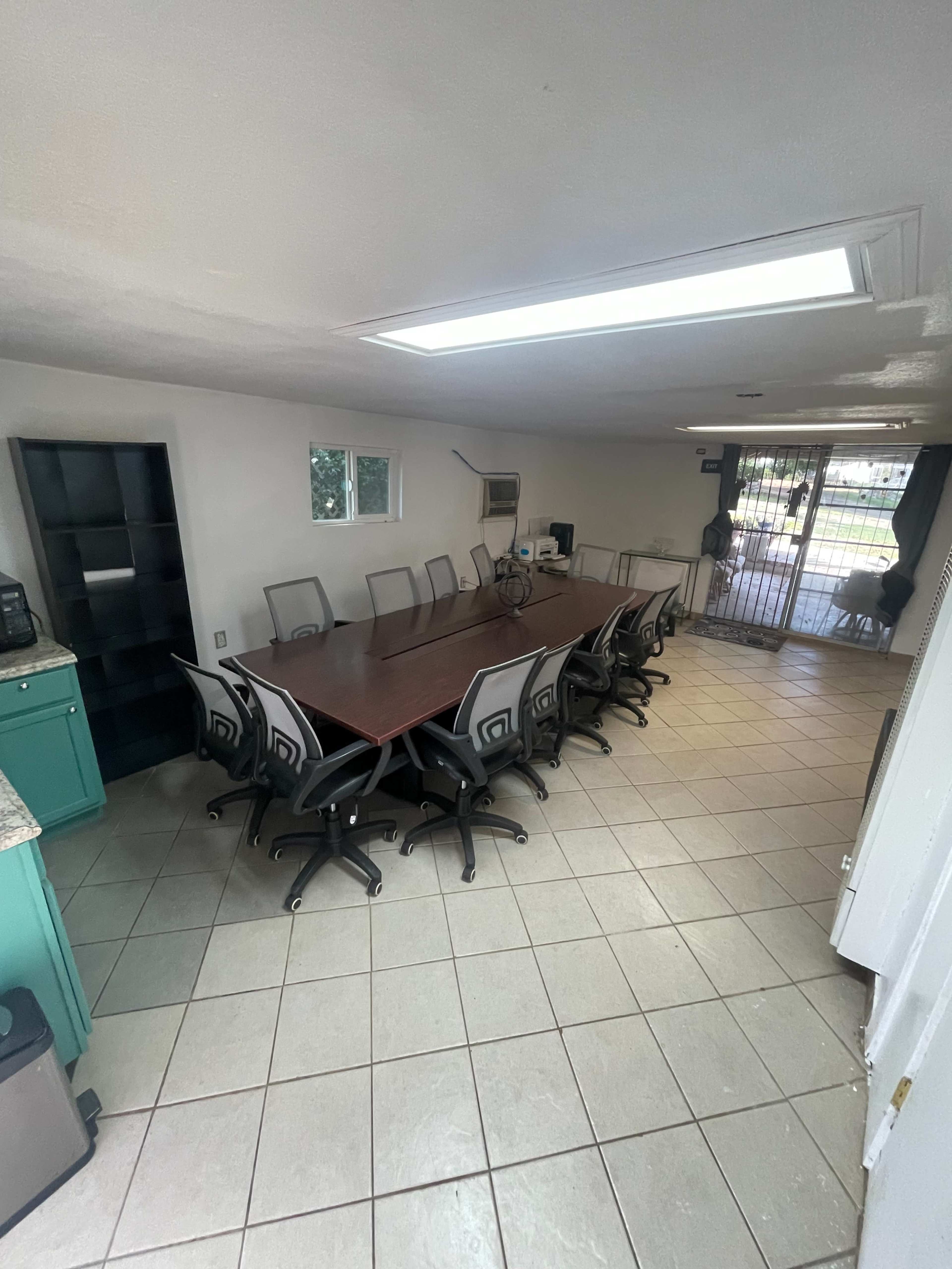 The image shows a conference room with a long rectangular table surrounded by multiple office chairs, tiled flooring, and a door leading outside.