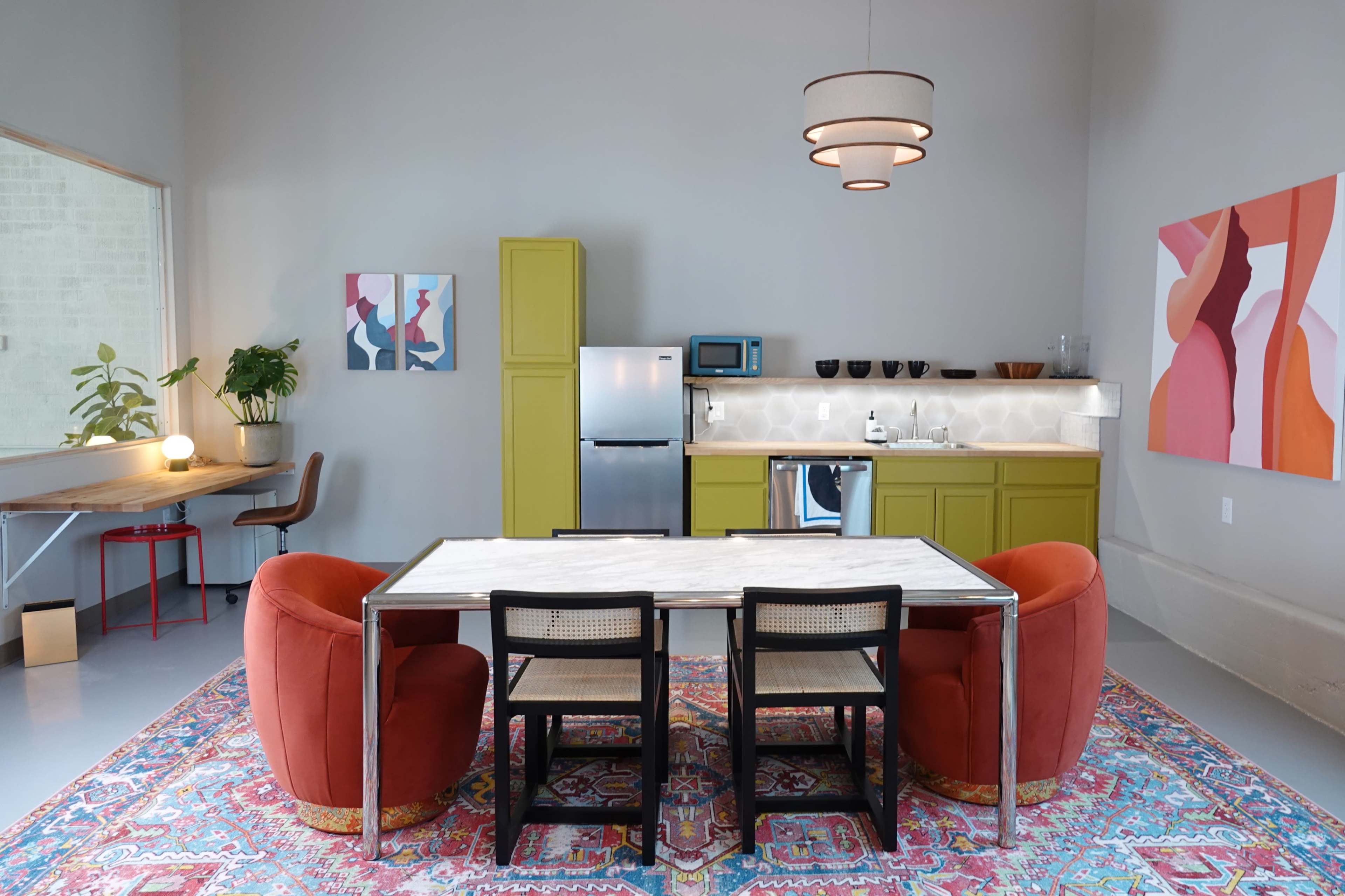 A modern kitchen and dining area features a marble-top table surrounded by black chairs, flanked by two red armchairs, with olive green cabinets and colorful artwork on the walls.