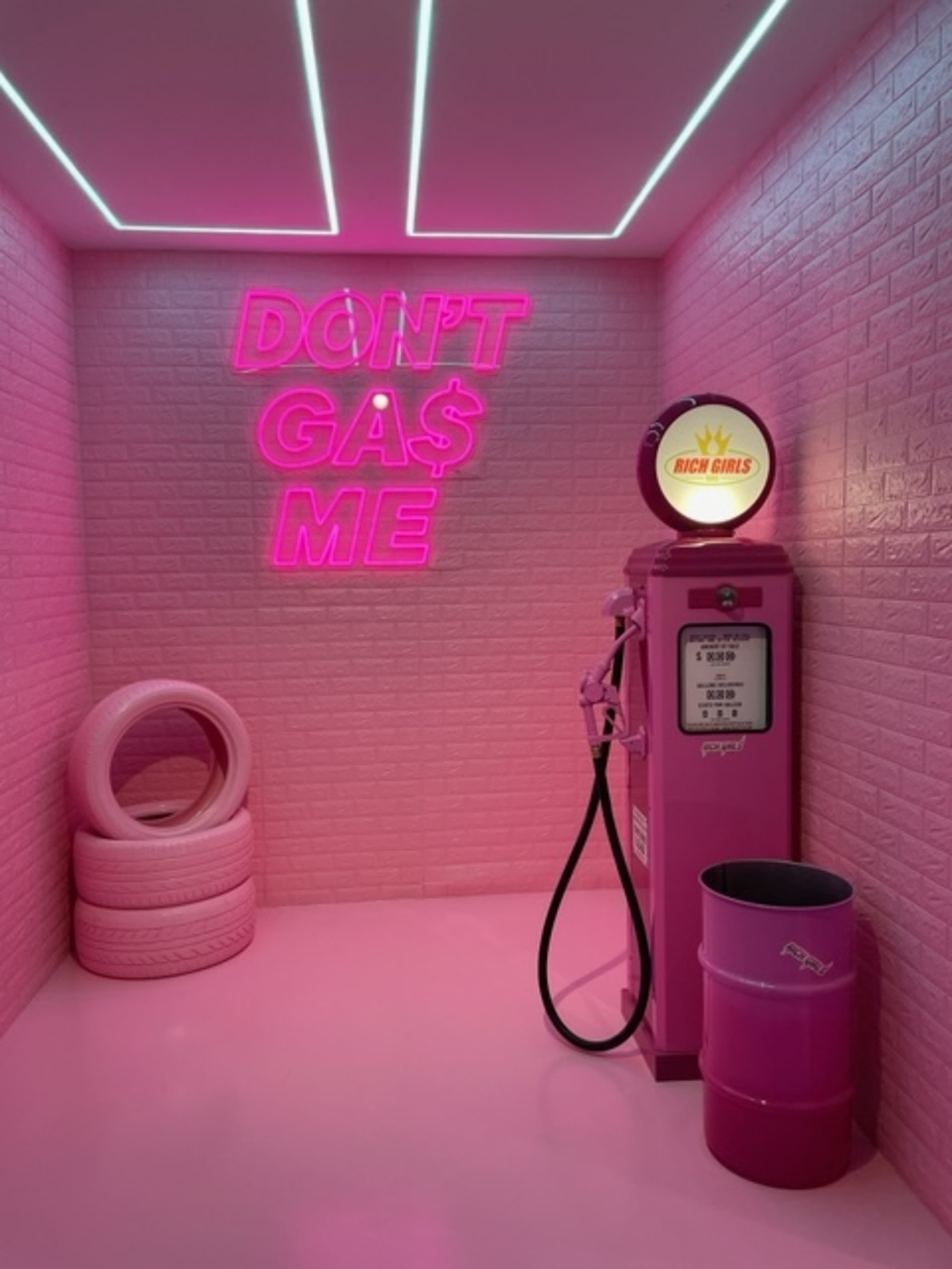 A pink-themed room featuring a vintage-style gas pump, neon signs, and pink tires against a backdrop of light-colored brick walls.