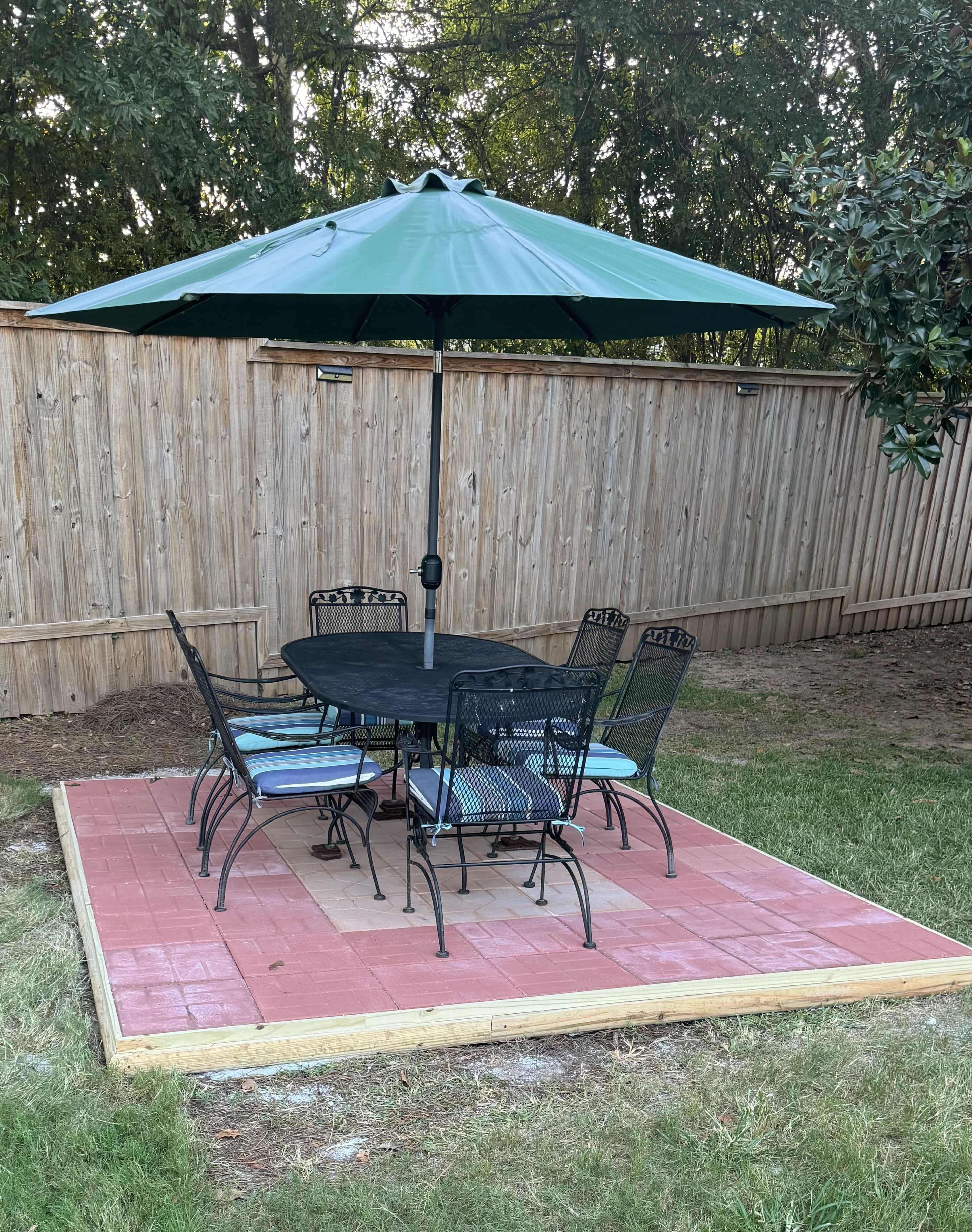 A rectangular, brick-paved patio features a black metal table with six matching chairs and a large green umbrella in a fenced backyard.