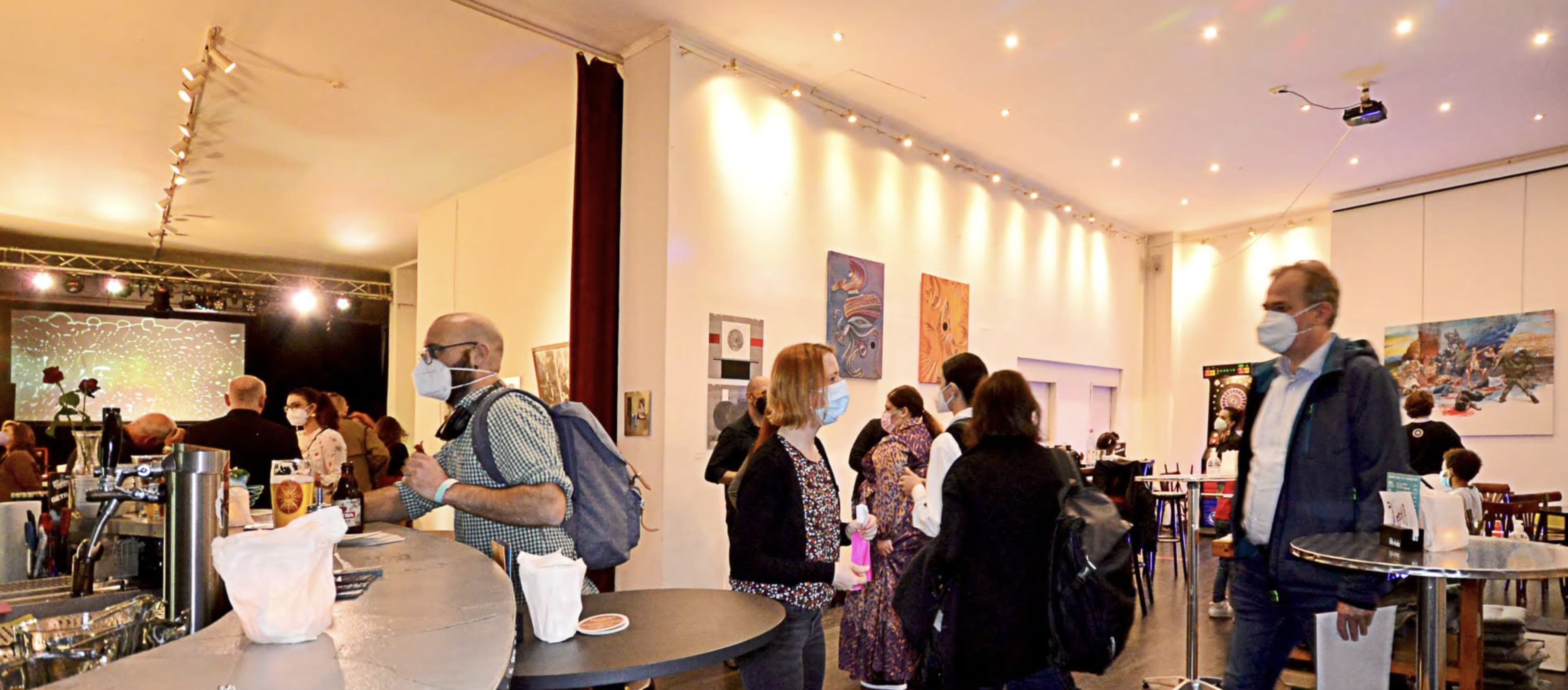 A crowded indoor space with people wearing masks, engaging in conversation and surrounded by art displayed on the walls.