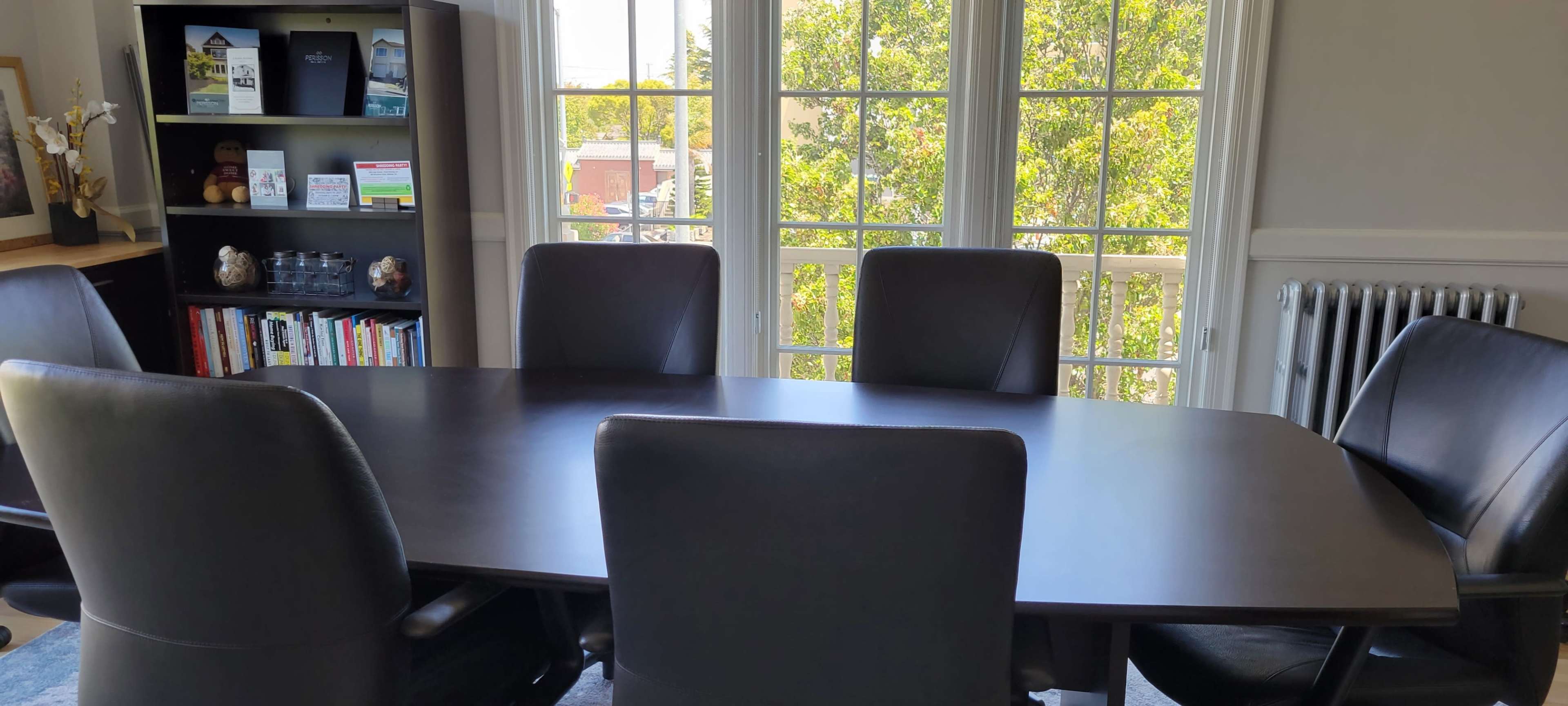 A conference room features a large, oval table surrounded by six dark chairs, with large windows providing a view of greenery outside.