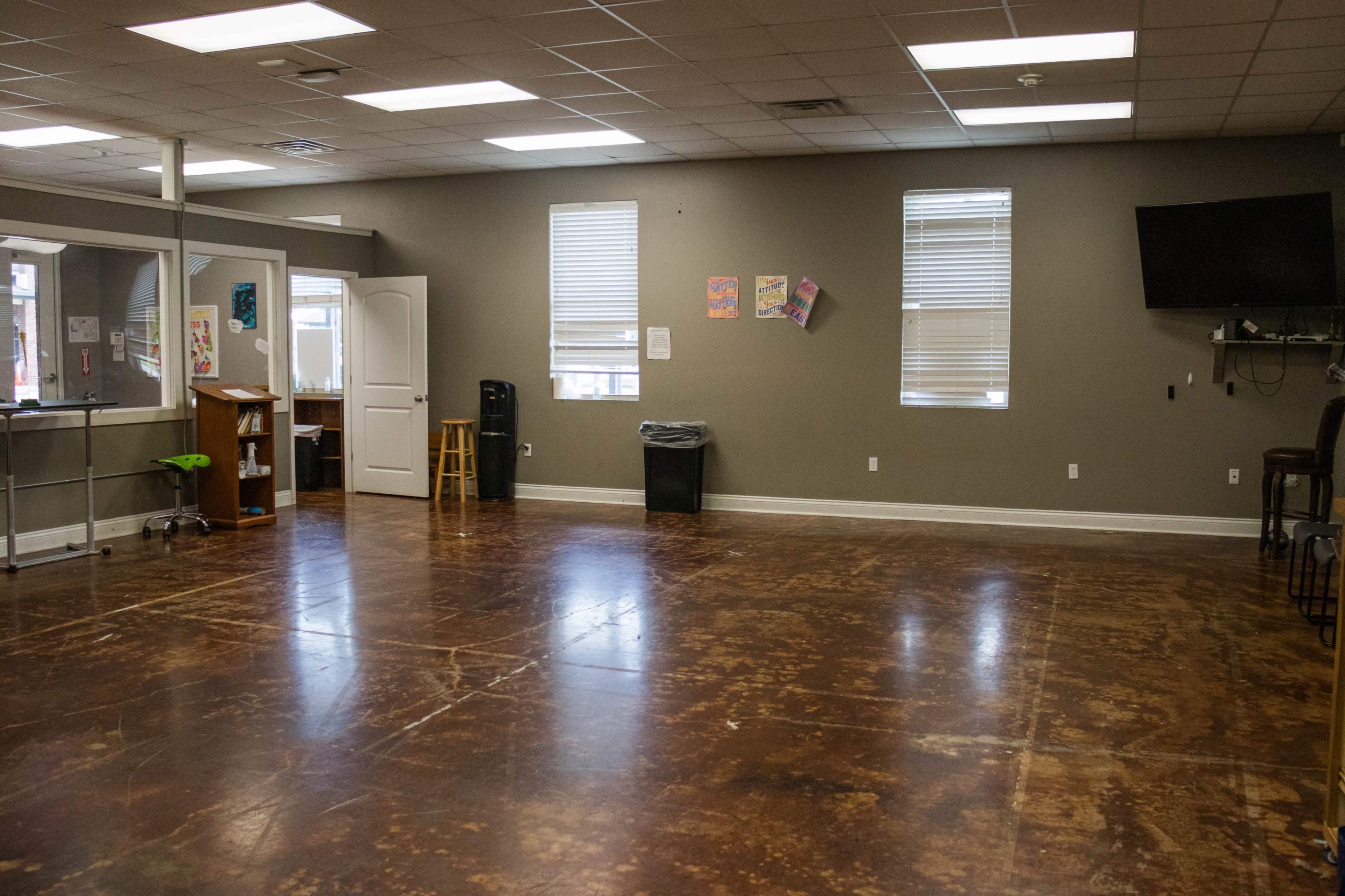 The image shows a spacious room with polished concrete flooring, a few windows, and minimal furniture including a stool and a trash can.