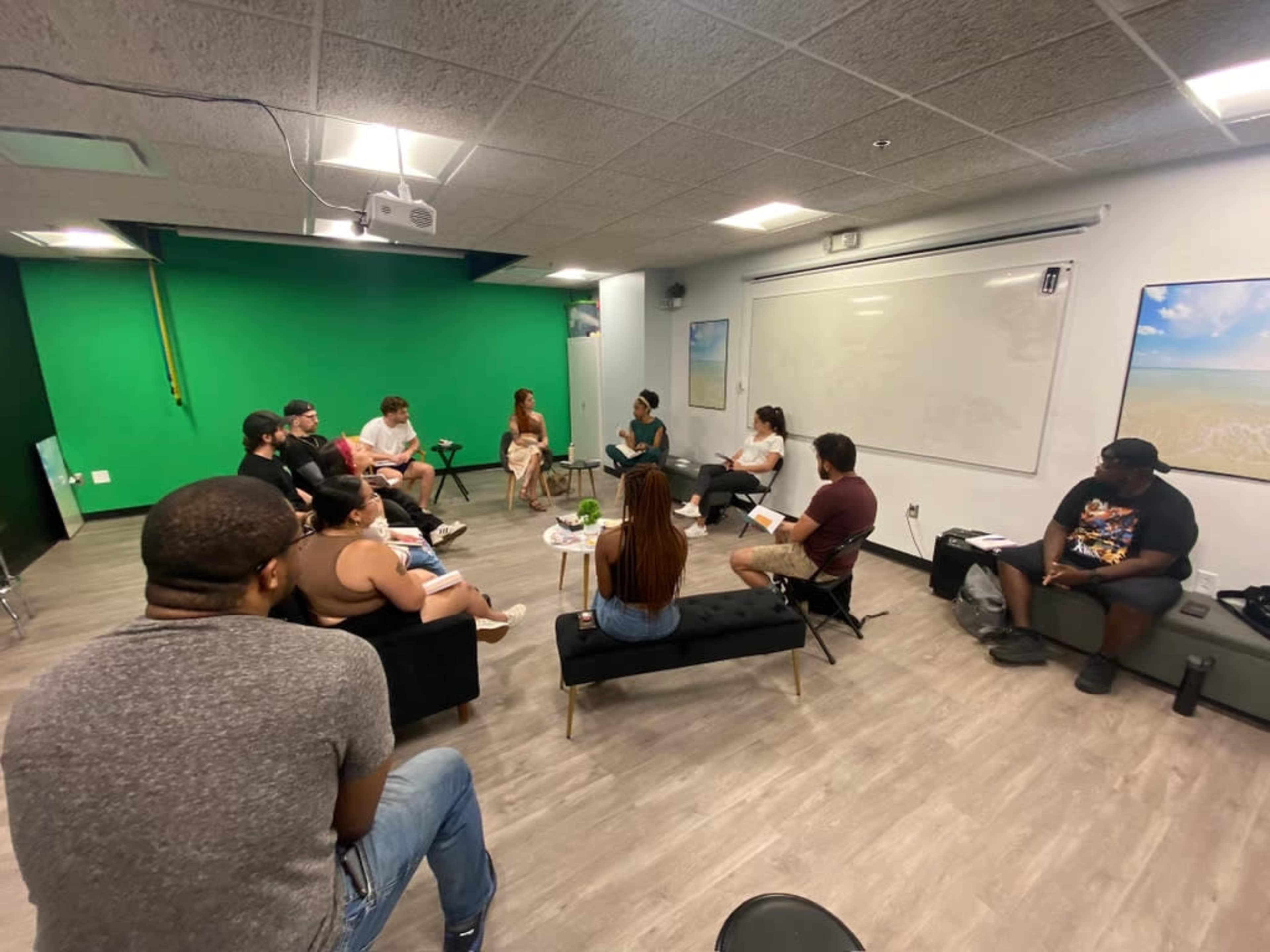 A group of people is gathered in a room with green walls and a projector, seated for a discussion or meeting.