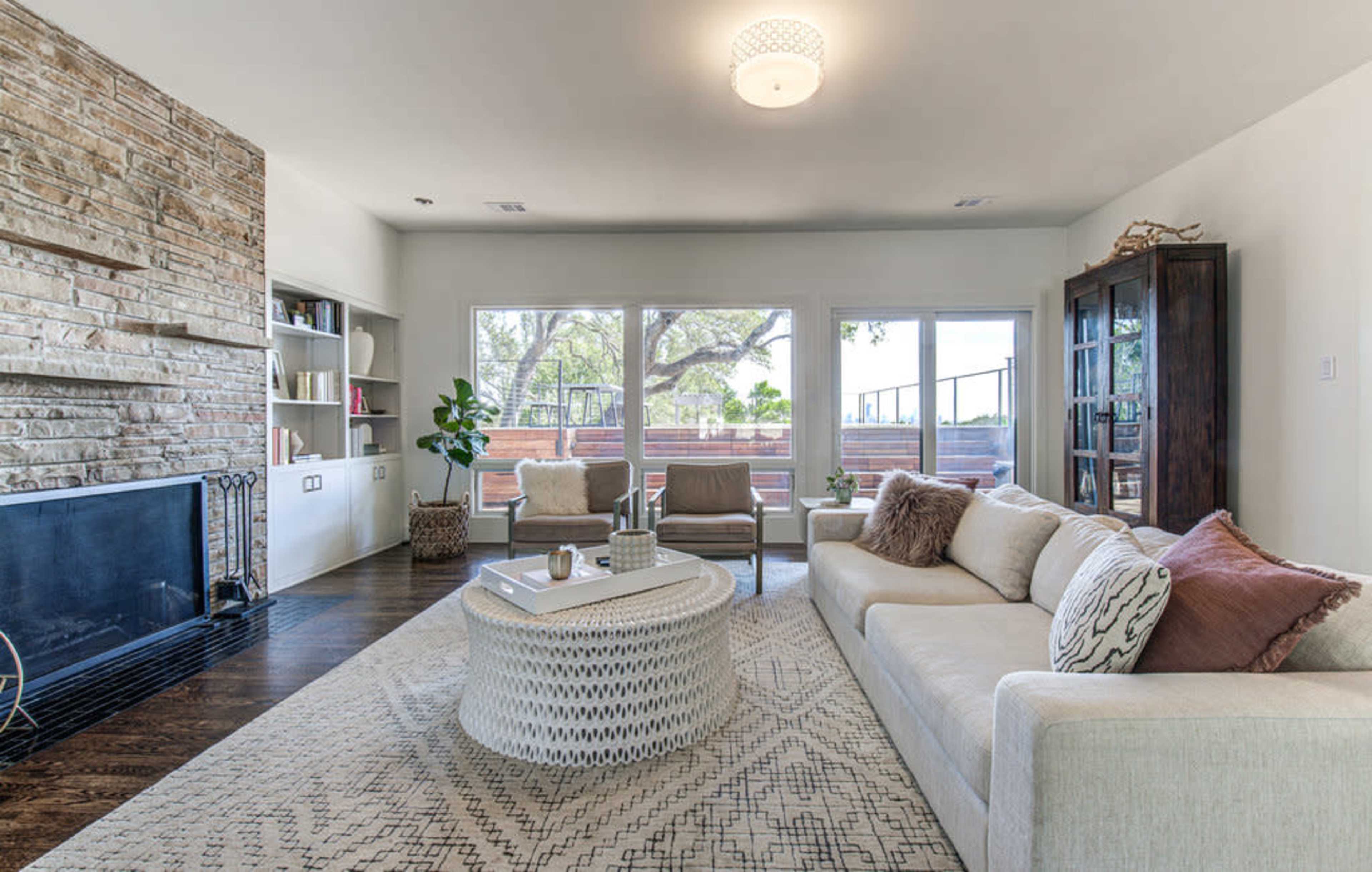 The living room features a modern design with a stone fireplace, large windows, and a circular coffee table surrounded by a sofa and armchairs.