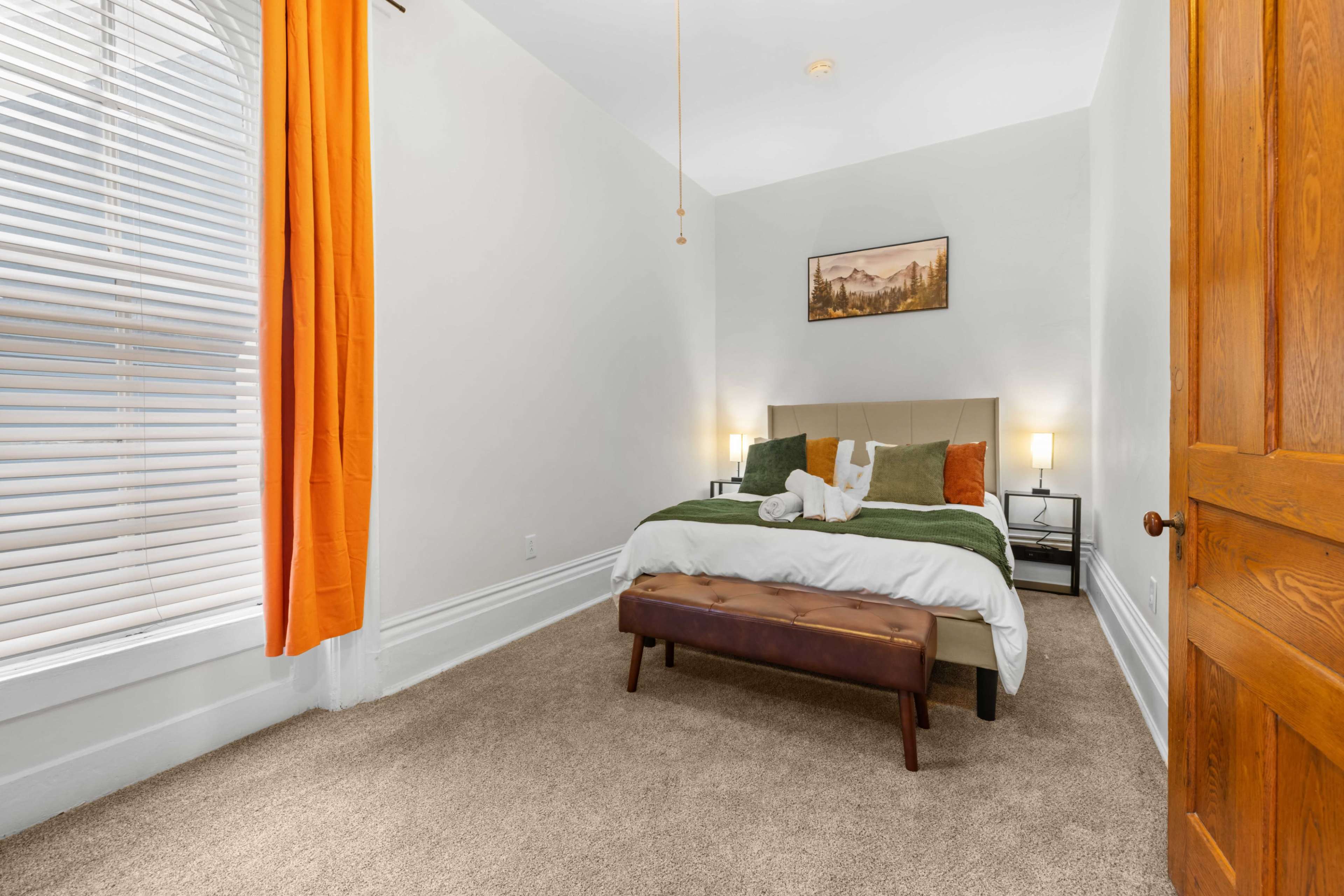 A neatly arranged bedroom features a bed with green and white bedding, a brown bench at the foot, and large windows with orange curtains.