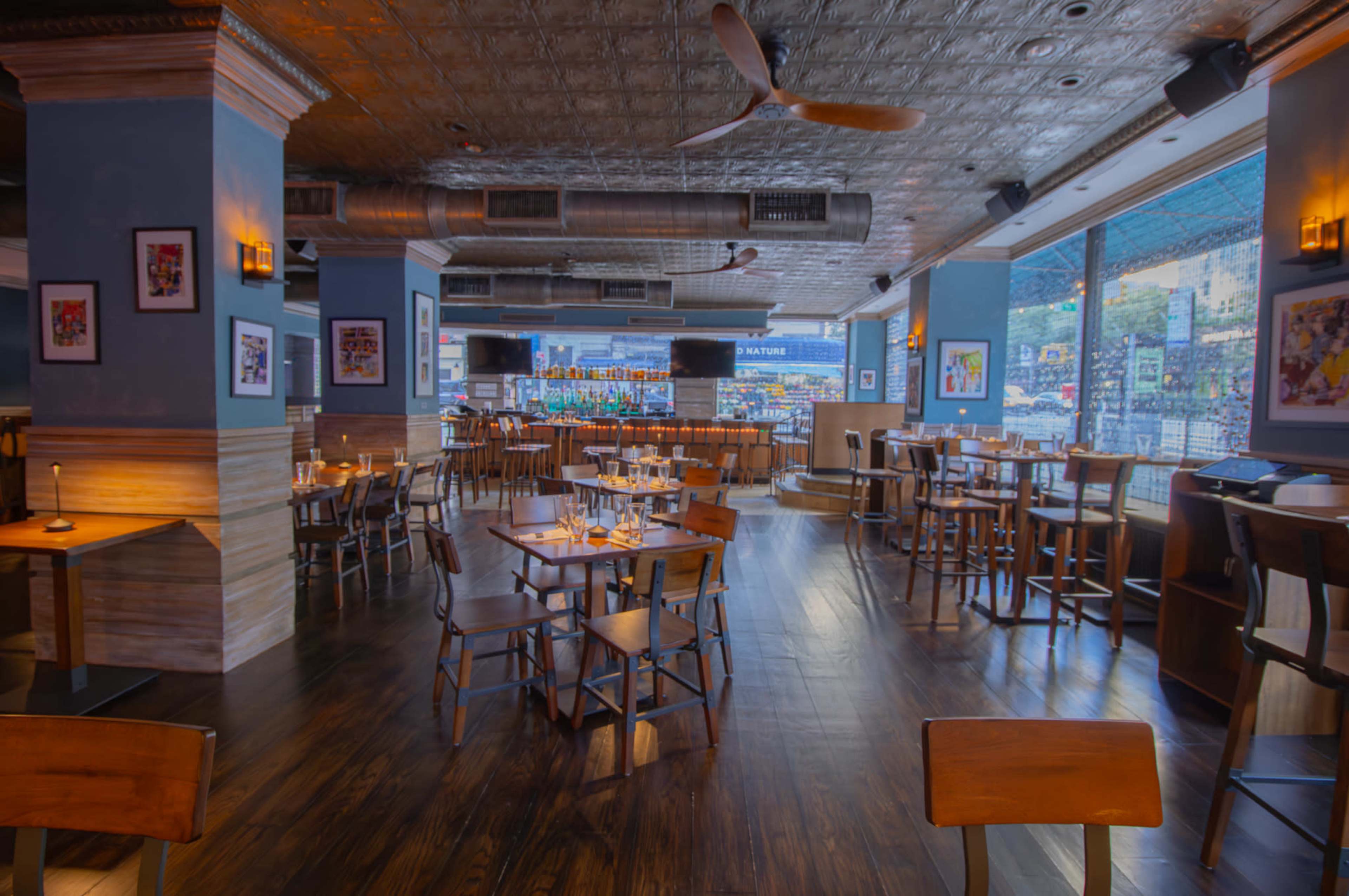 The image shows the interior of a restaurant with wooden tables and chairs, large windows, and a bar area in the background.