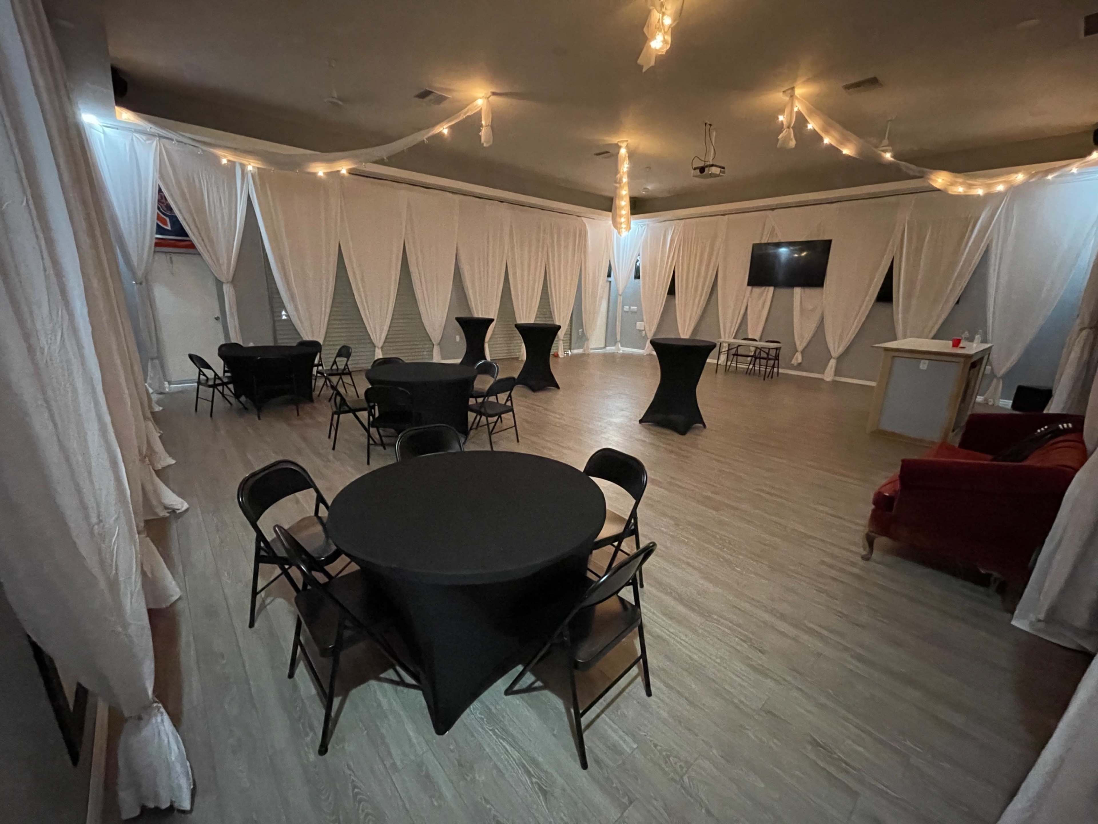 The image shows a spacious event room with several round black tables and chairs, white draped curtains, and soft lighting.
