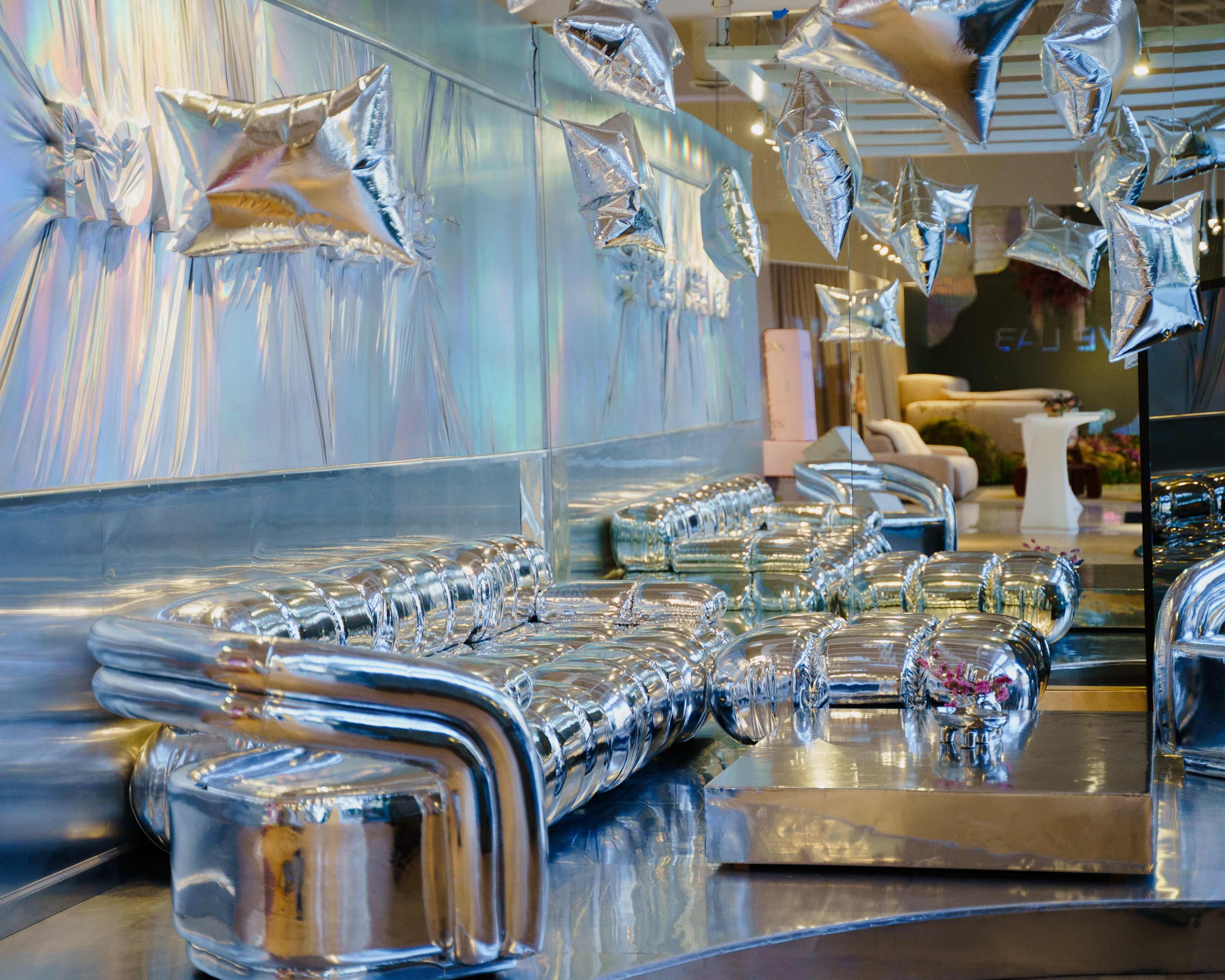 The image features a modern, reflective lounge area with metallic furniture and shiny, balloon-like decor suspended from the ceiling.