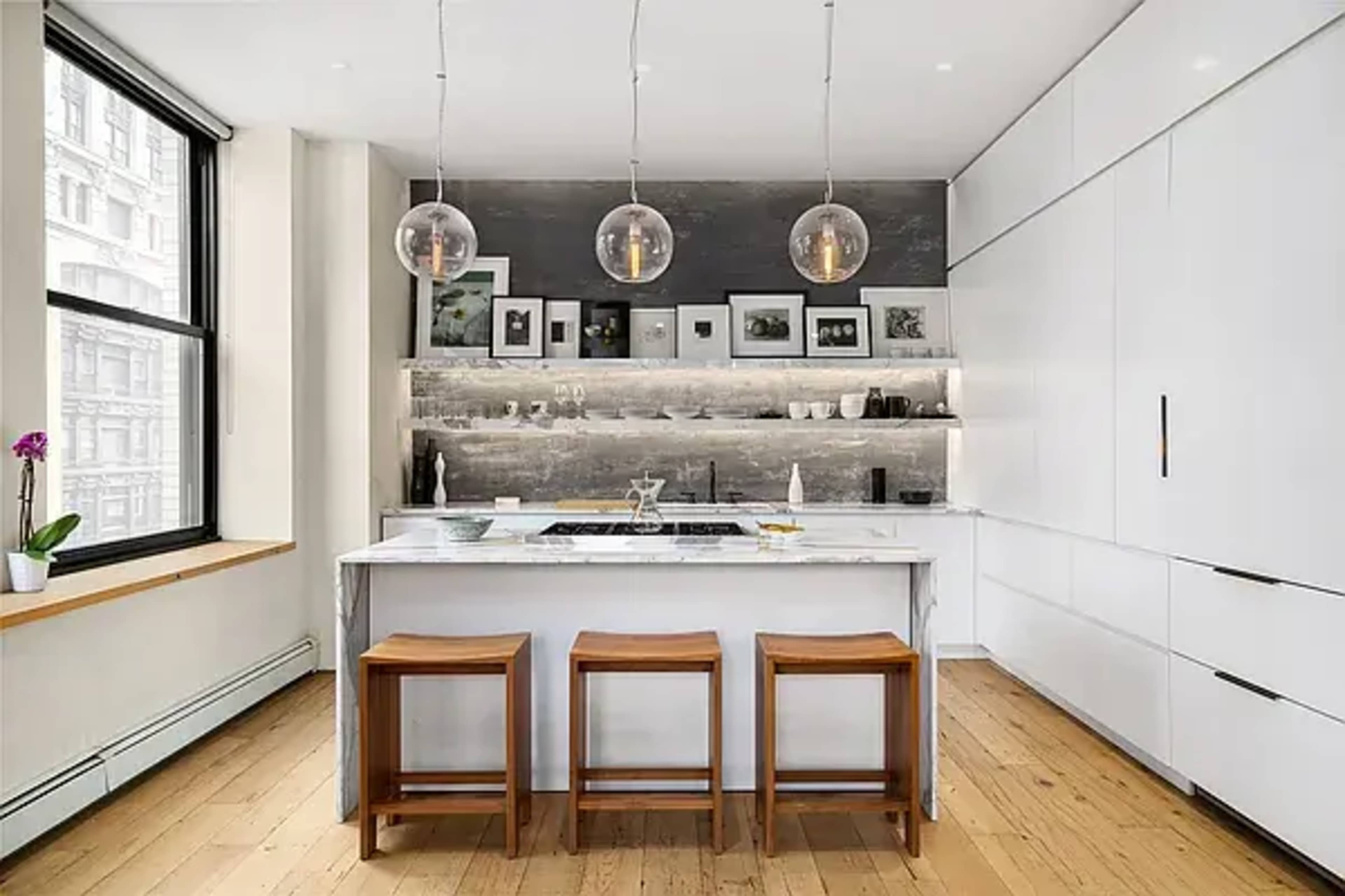 The kitchen features a marble island with three wooden stools, pendant lights overhead, and a backdrop of shelves containing various dishware and framed photographs.