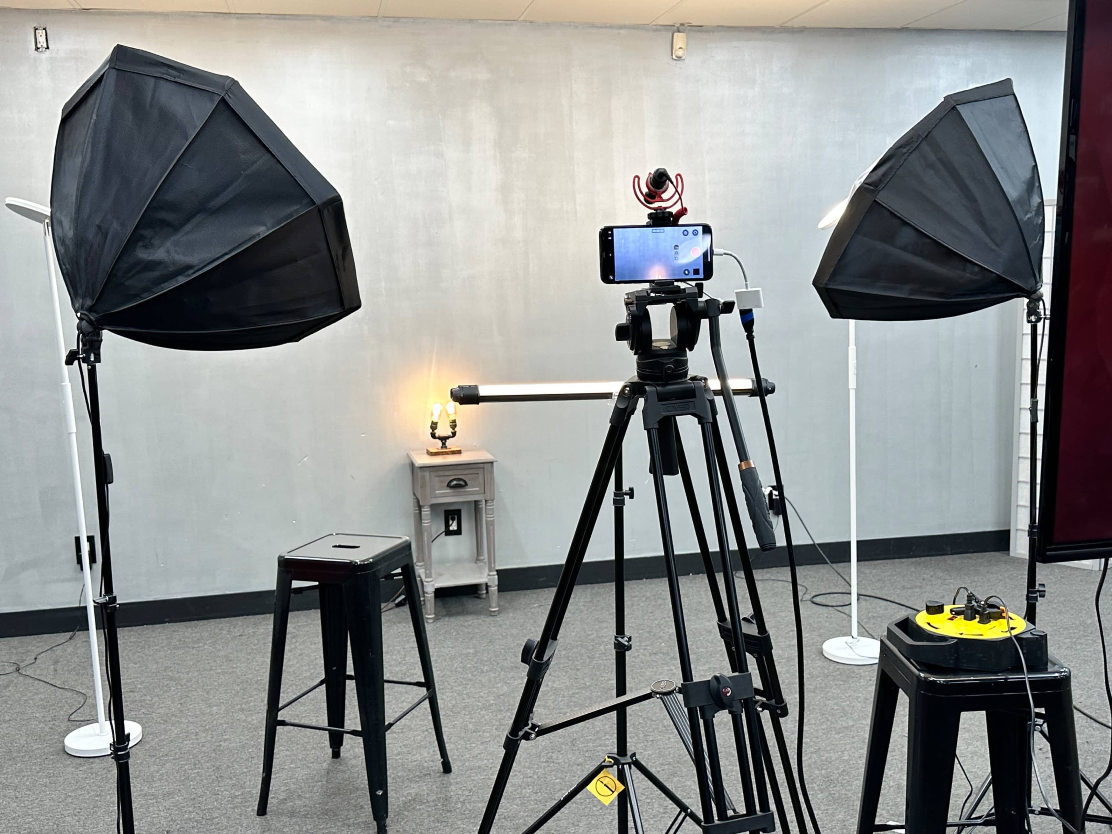A photography setup with two softbox lights, a smartphone mounted on a tripod, a stool, and a small table with a lamp in a room with gray walls.