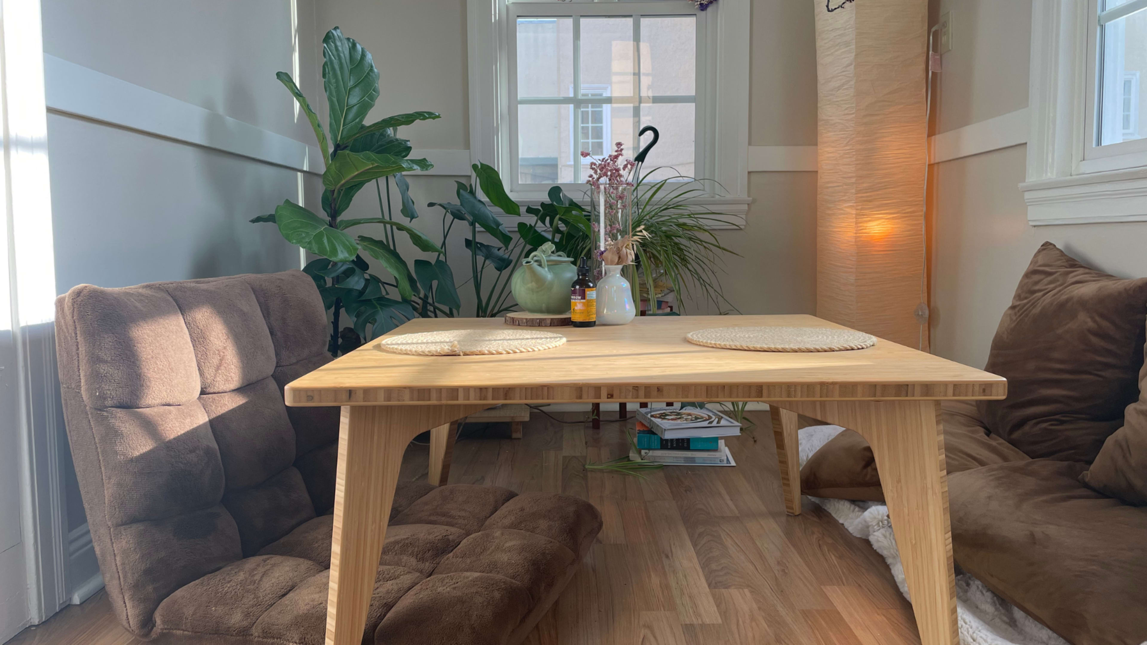 A wooden table with two placemats is set between two brown floor cushions, surrounded by plants and soft lighting.