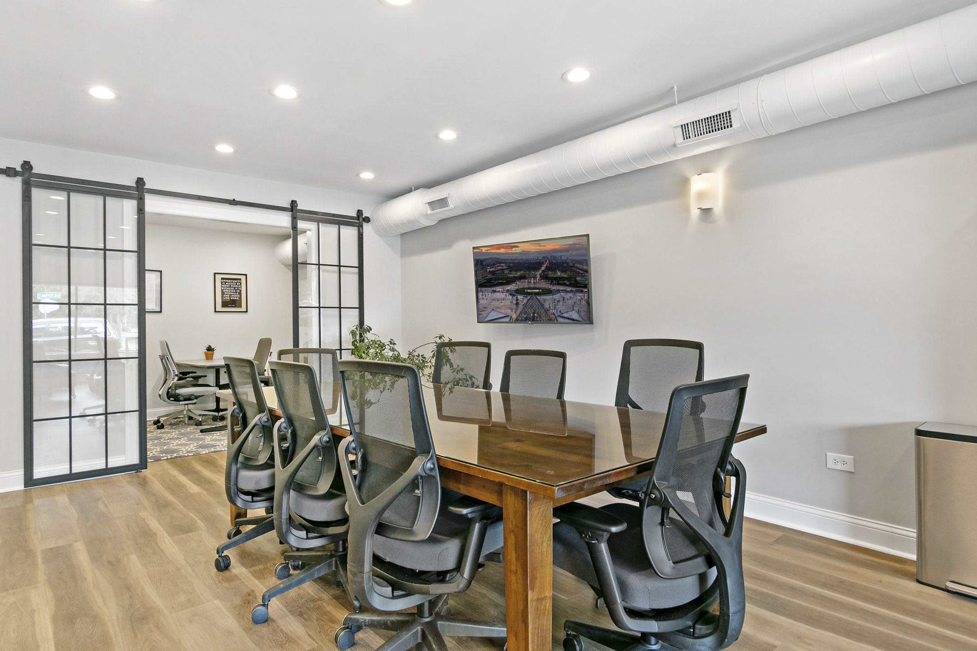 A modern conference room features a long wooden table surrounded by black mesh chairs, with glass-paneled doors and wall-mounted artwork.