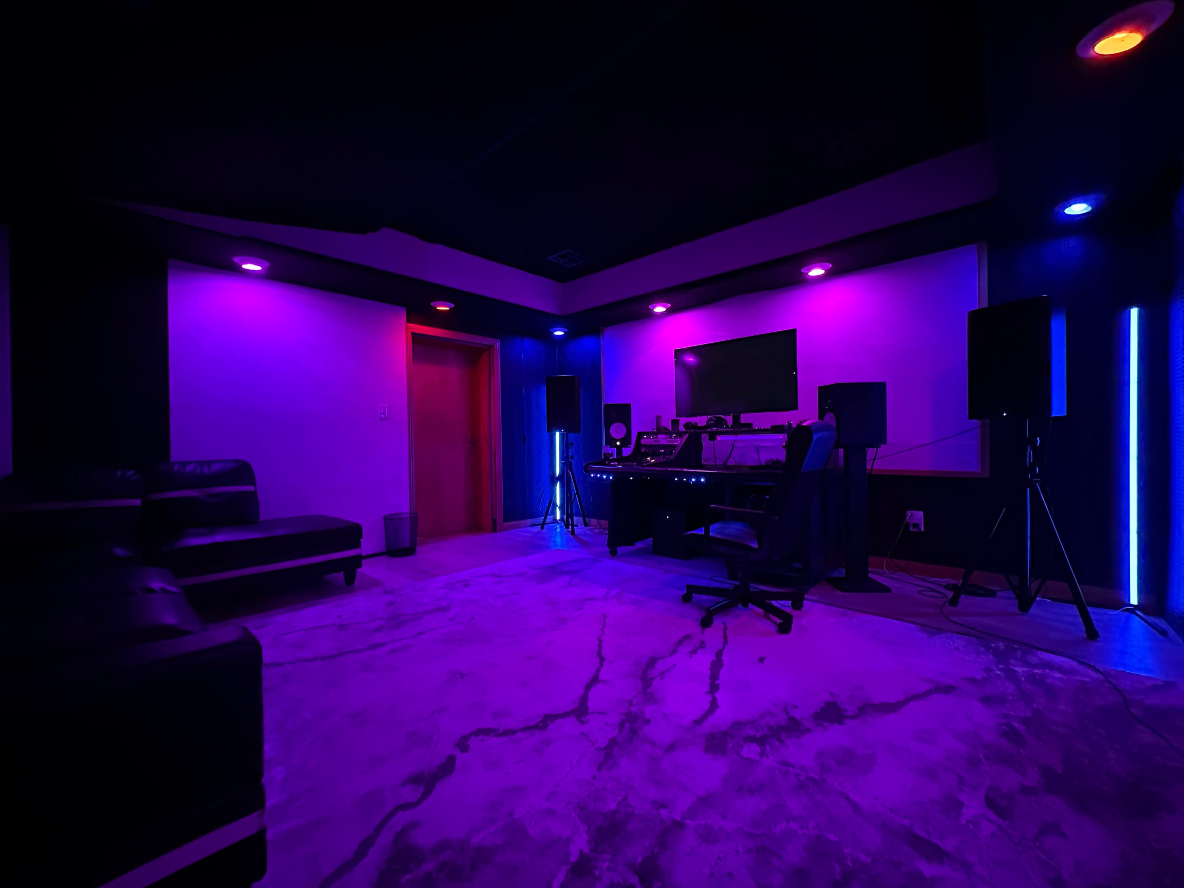 A modern recording studio with purple and blue lighting, featuring a desk with audio equipment, a chair, and a comfortable couch.