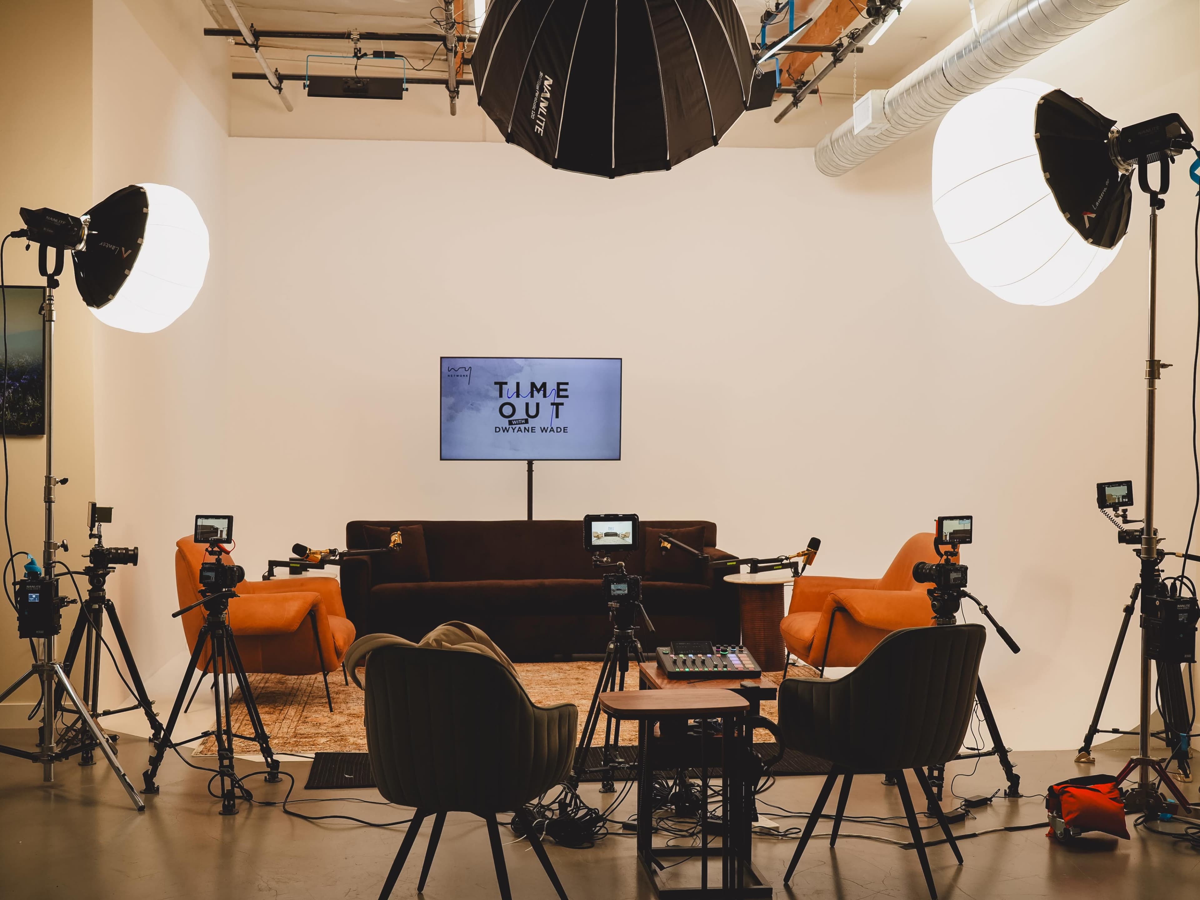 A well-equipped studio setup features a large screen displaying "TIME OUT," a central sofa, two chairs, and multiple cameras and lighting equipment arranged for a recording session.