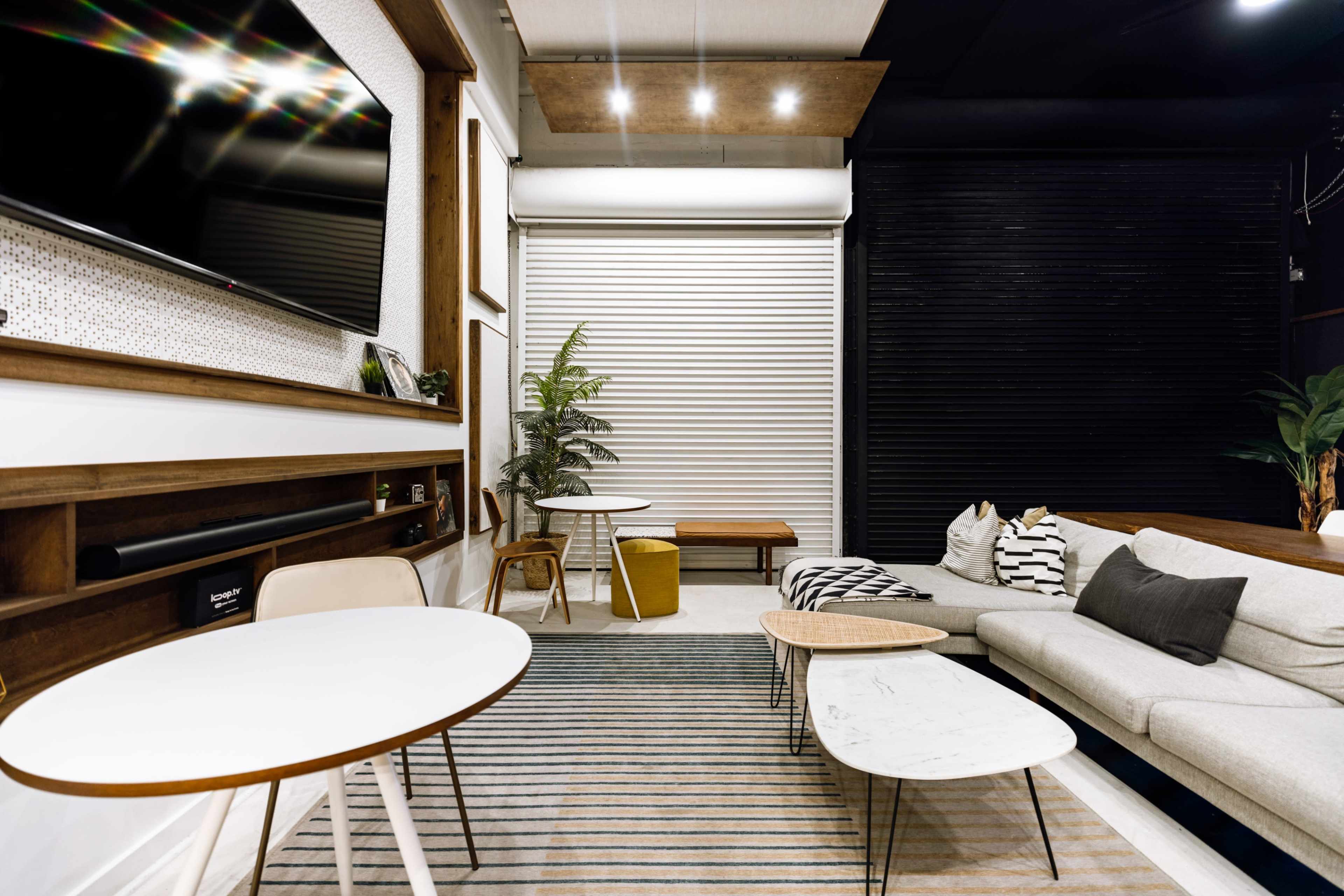 The image shows a modern lounge area featuring a sectional sofa, two coffee tables, potted plants, and a wall-mounted TV against a backdrop of contrasting dark and light surfaces.