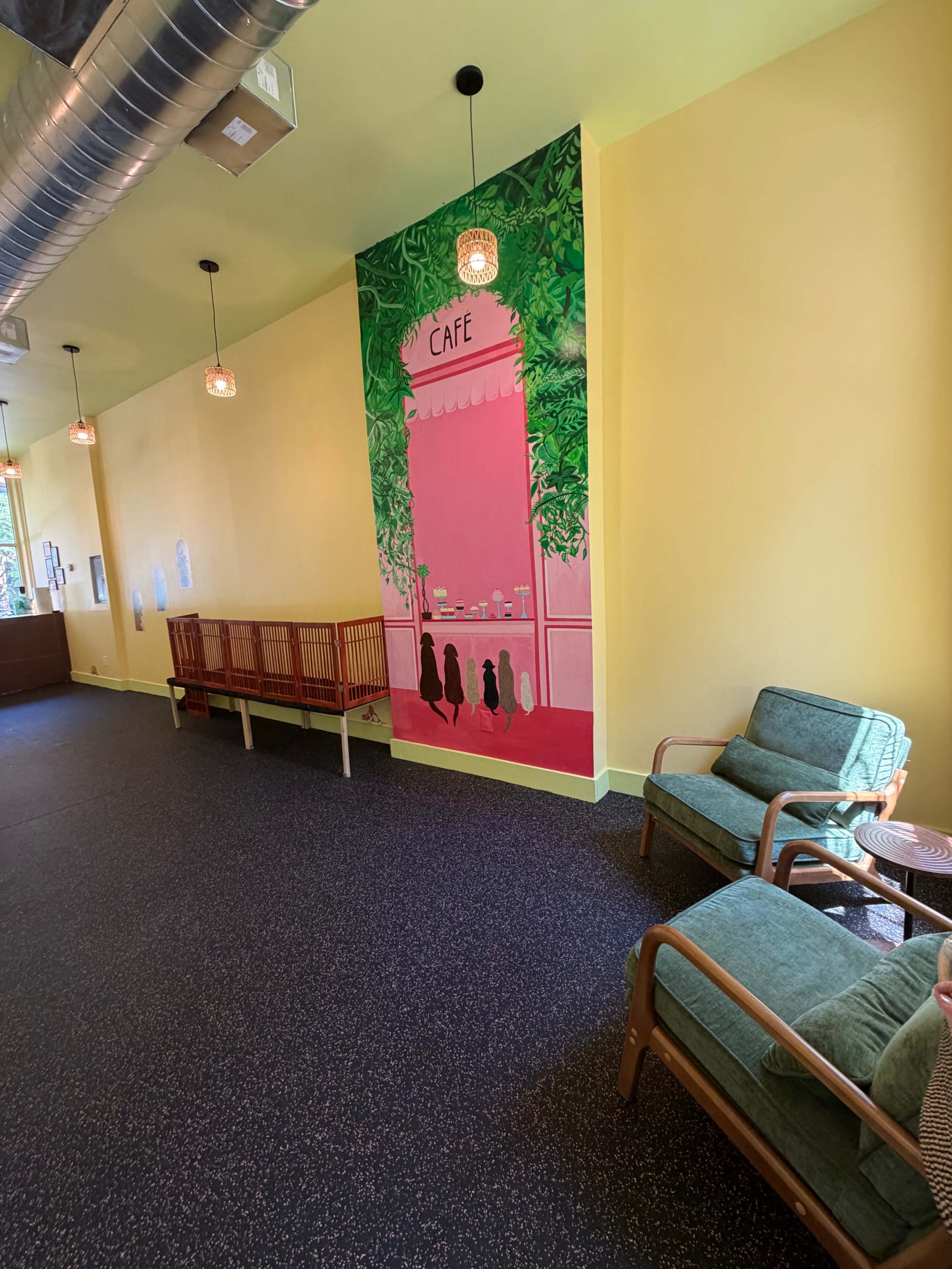 The image shows a brightly colored café interior with a mural on the wall, featuring a pink café scene surrounded by green foliage, and simple seating arrangements in the foreground.