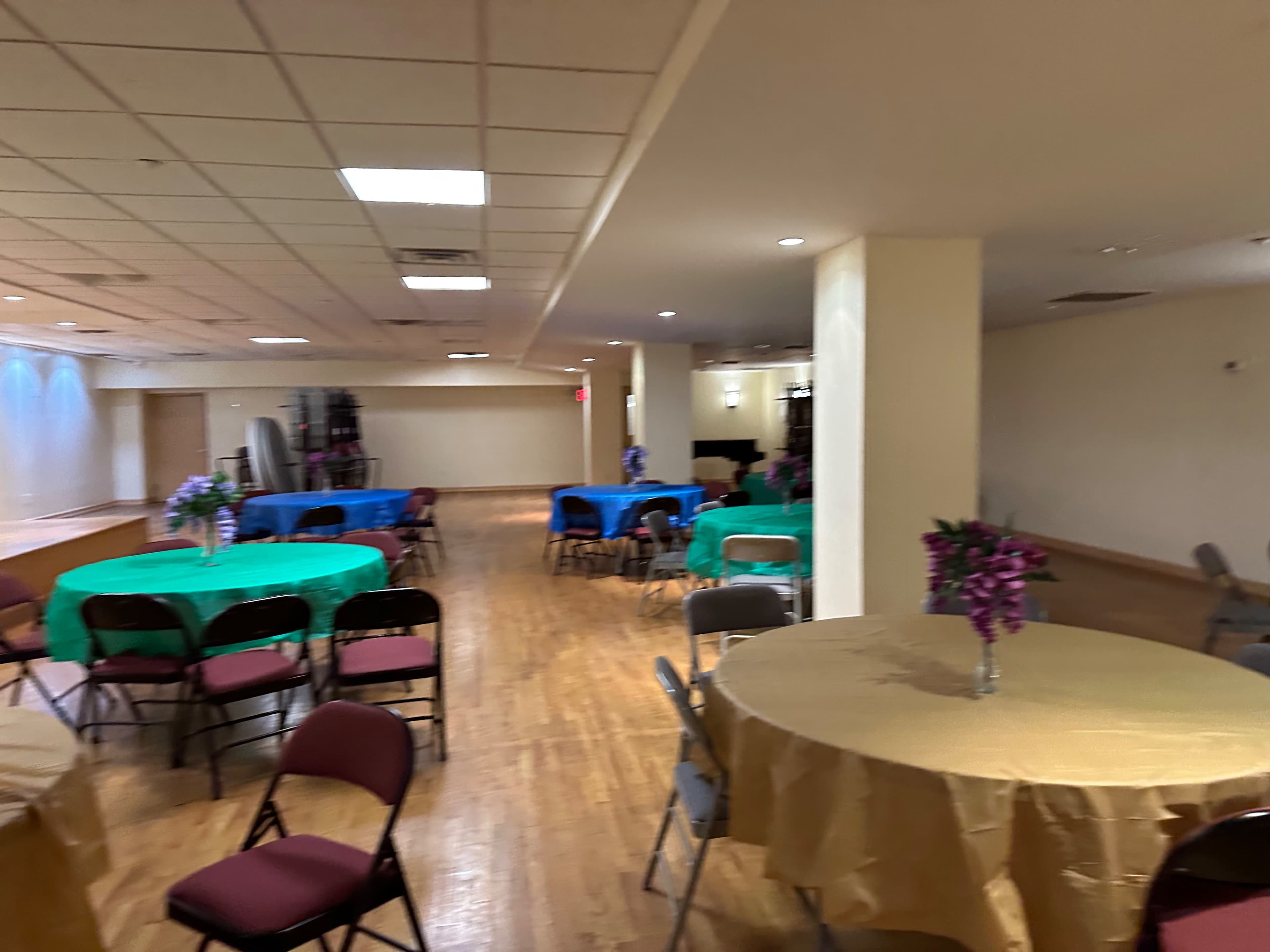A spacious indoor venue features several round tables covered with colorful tablecloths and arranged in rows along a wooden floor.