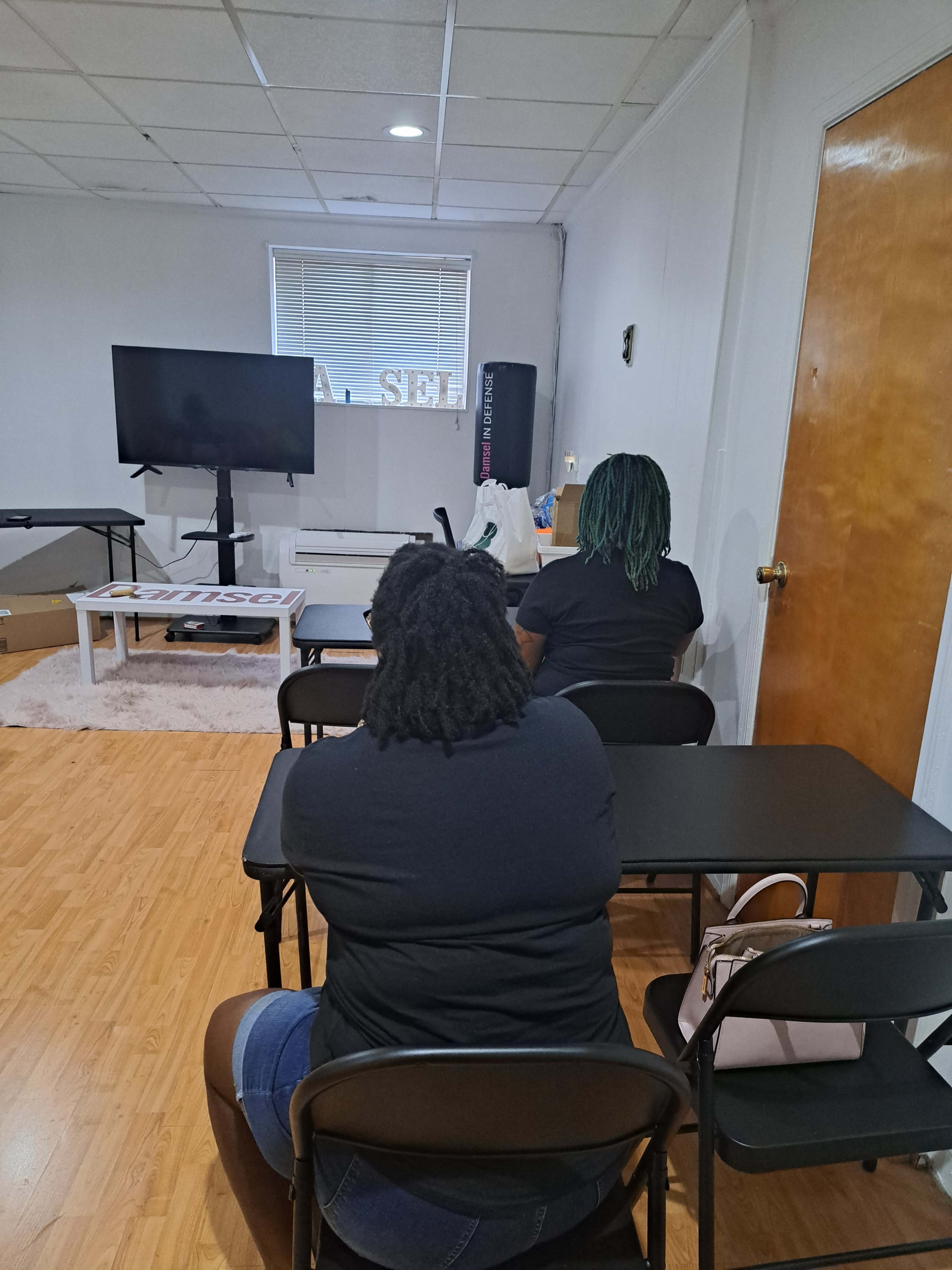 Two people sit at black folding tables, facing a large television screen in a room with wooden flooring and white walls.